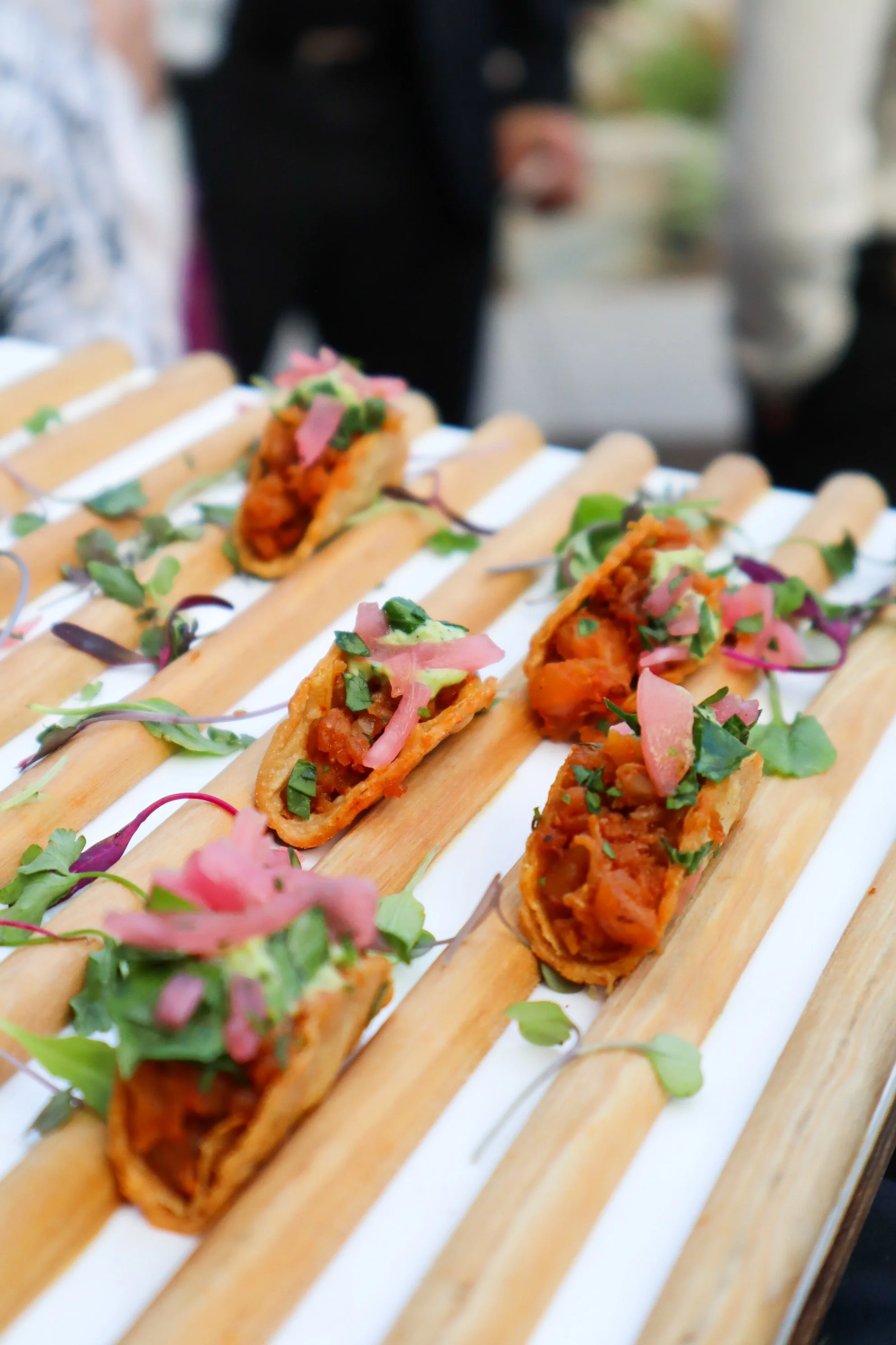 Close-up of three mini taco bites with meat, topped with chopped herbs, pickled onions, and microgreens, arranged on a wooden display.
