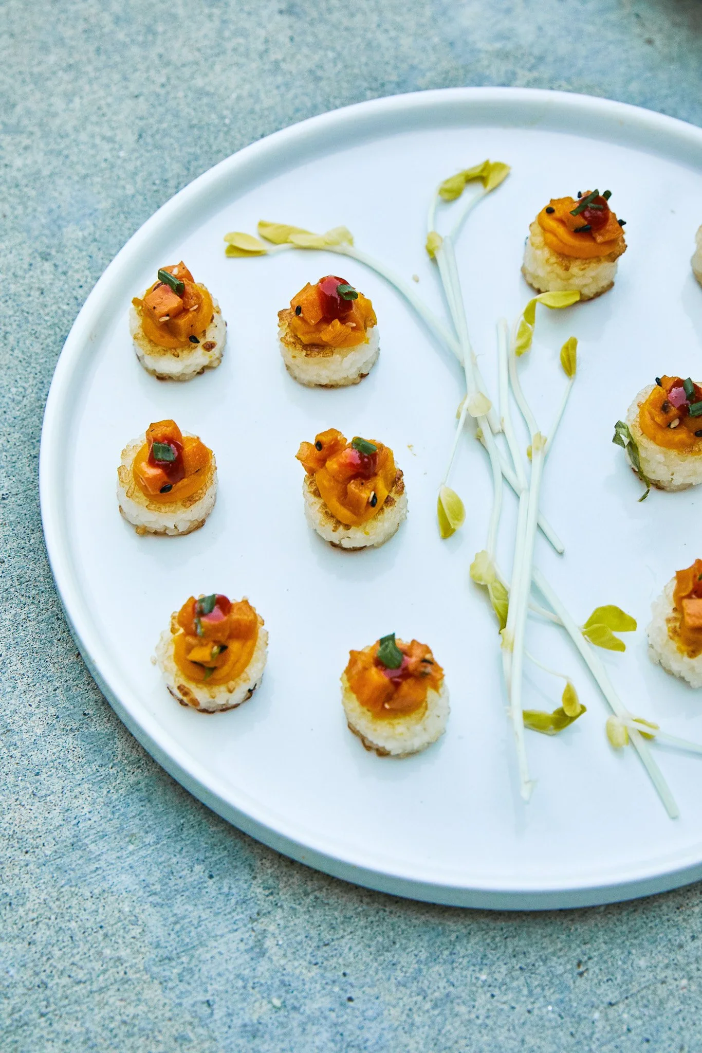 Assorted sushi bites topped with orange fish roe and garnished with chopped green herbs, arranged on a white plate with a few bean sprouts and green leaves.