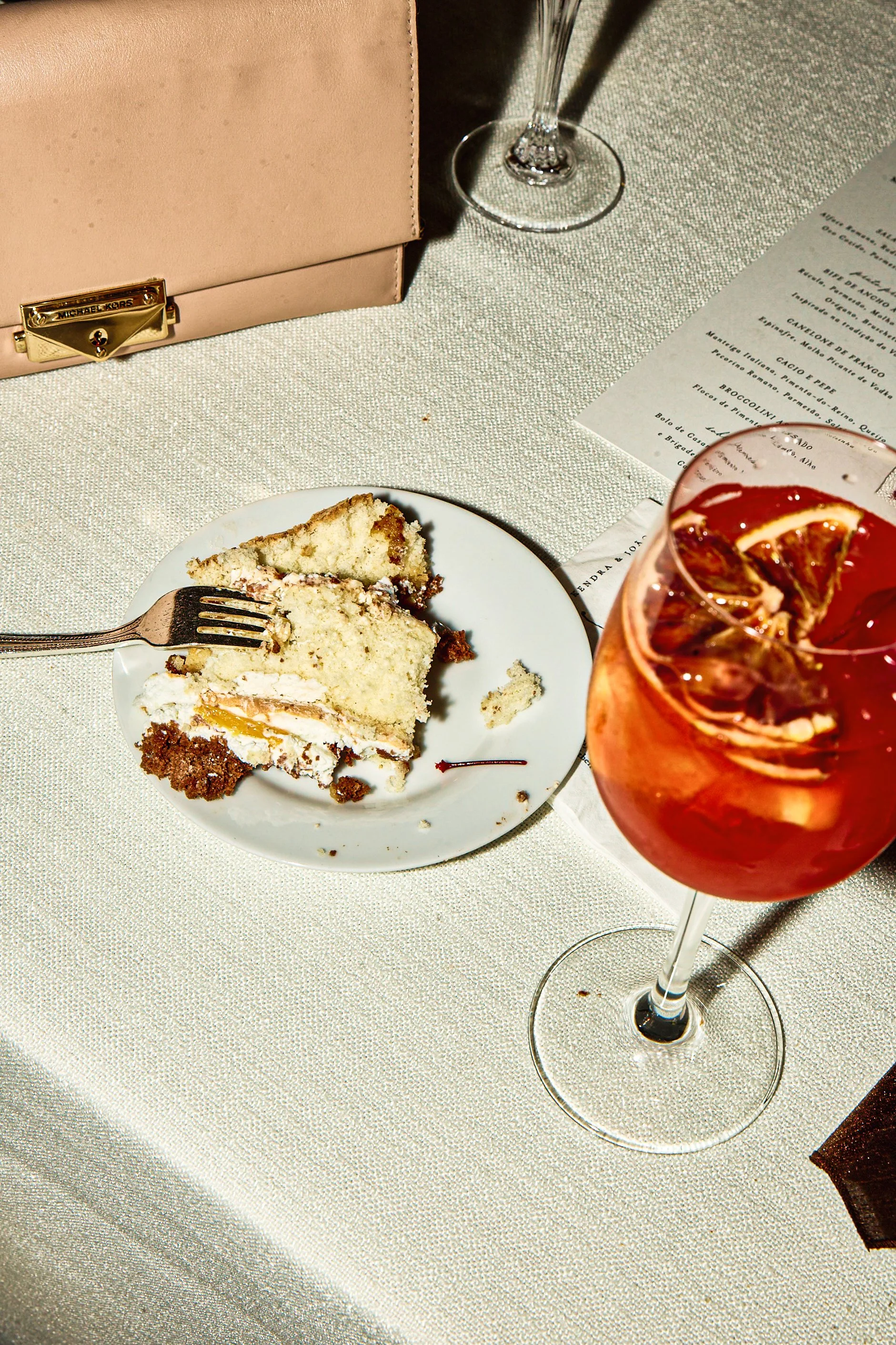 A partially eaten slice of cake on a white plate with a fork, a glass of pink cocktail with lime slices, a beige handbag, and a wine glass on a table with a menu and a textured tablecloth.