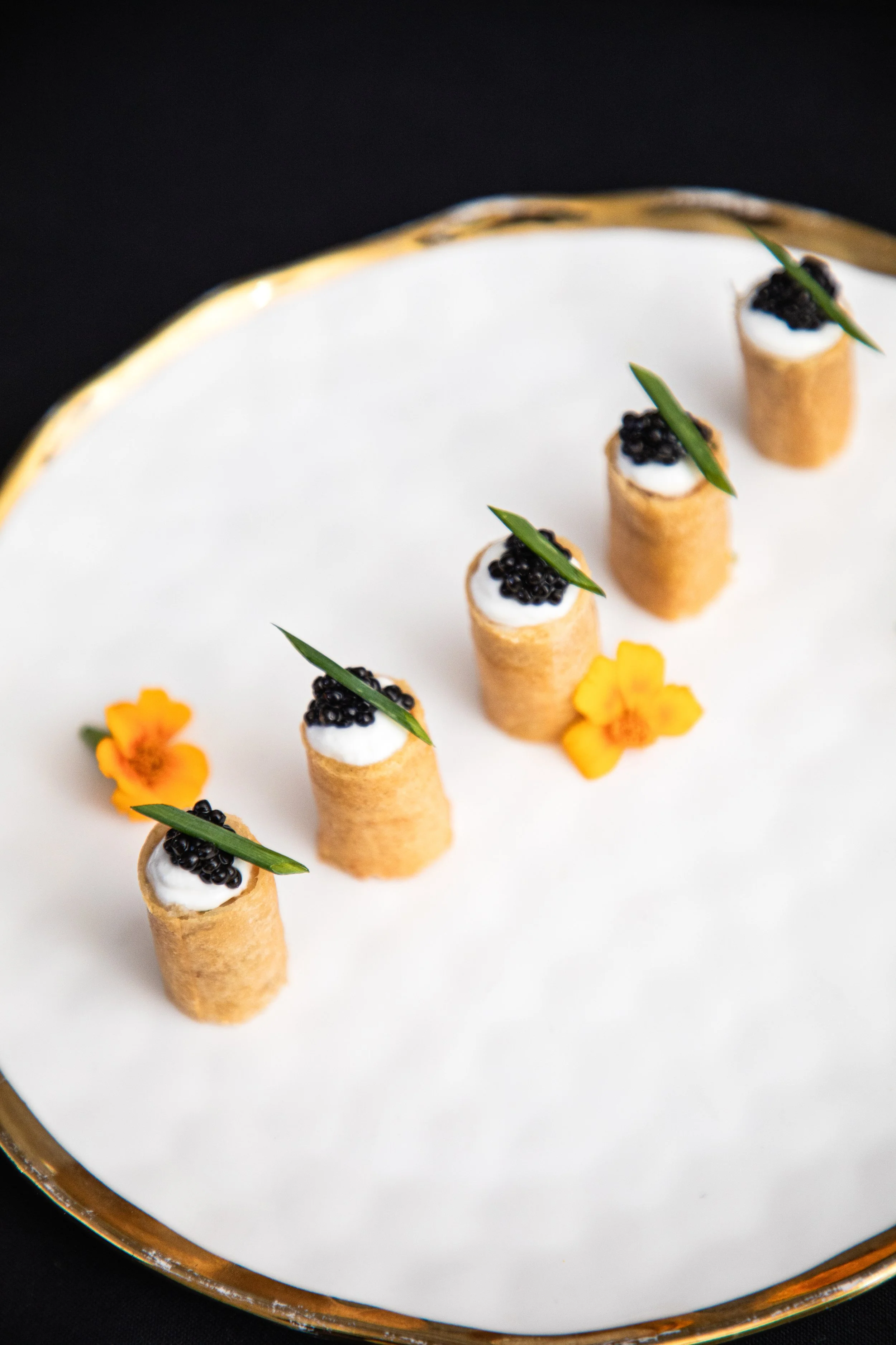 Five small appetizers with crispy shells, topped with black caviar, white cream, and garnished with small green leaves, arranged on a white plate with yellow flowers.