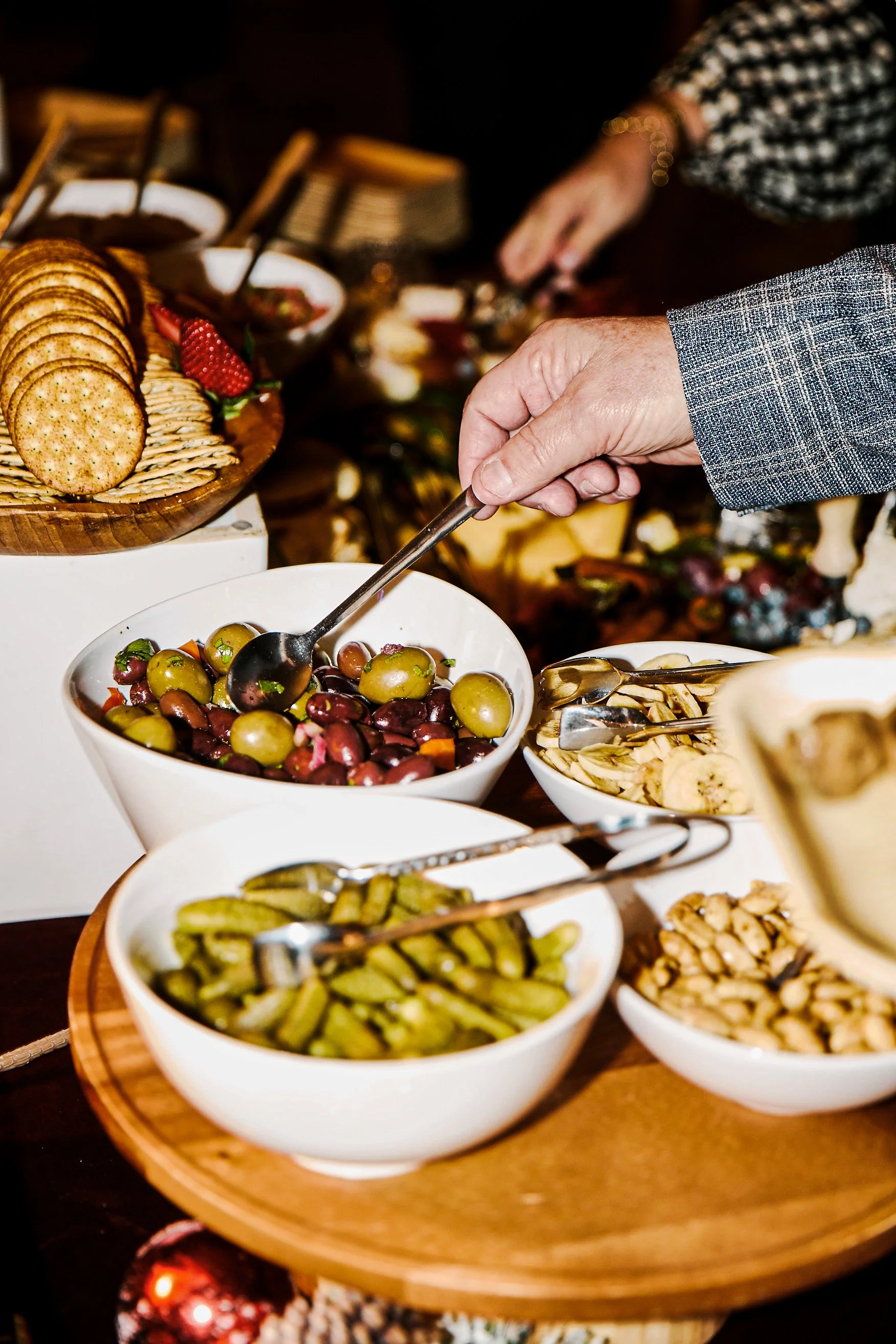 Close-up of a person in a gray suit grabbing green olives from a white bowl at a buffet table with bowls of pickles, grapes, and nuts, and crackers on a wooden platter in the background.