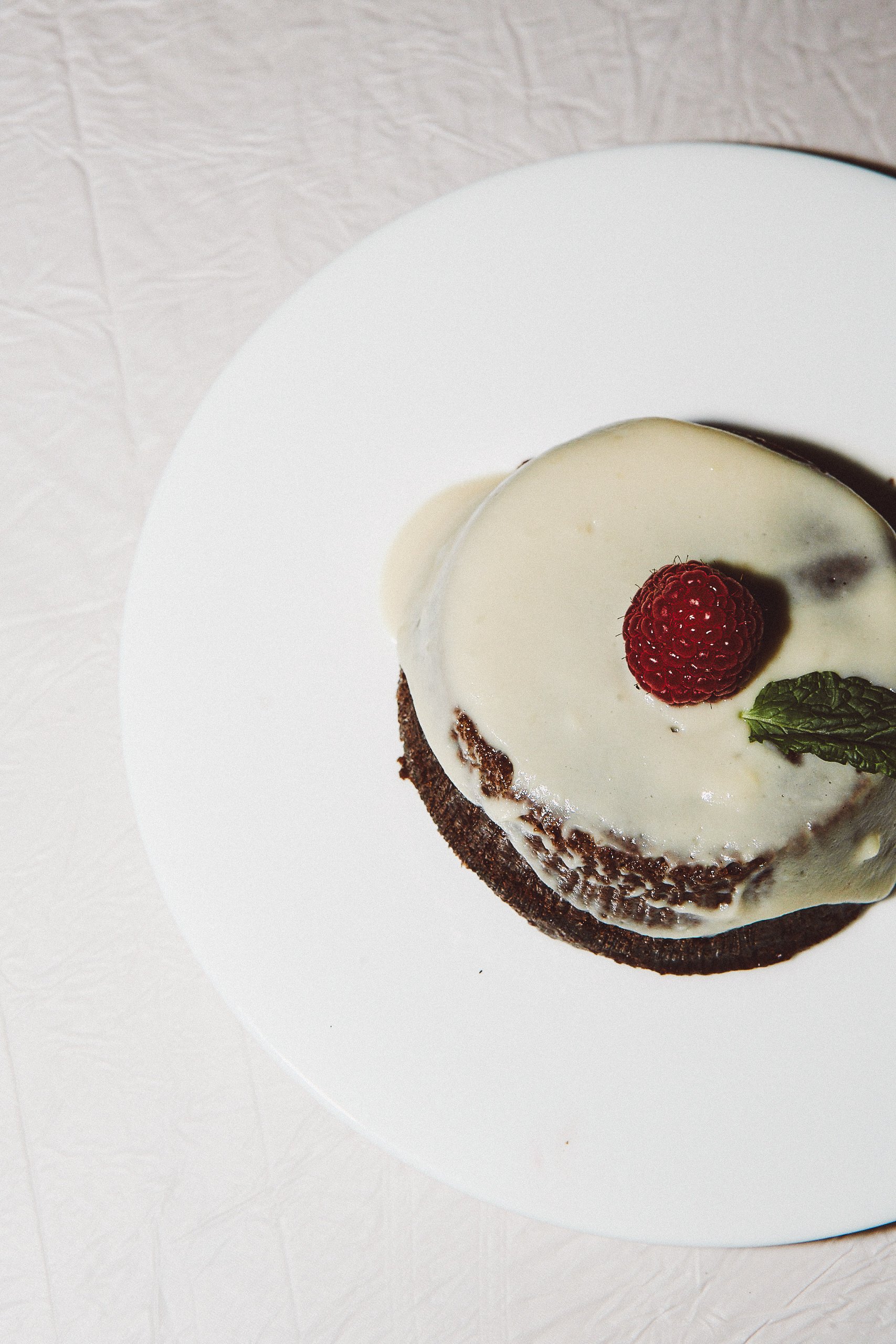 Chocolate cake with white frosting, topped with a raspberry and a mint leaf on a white plate.