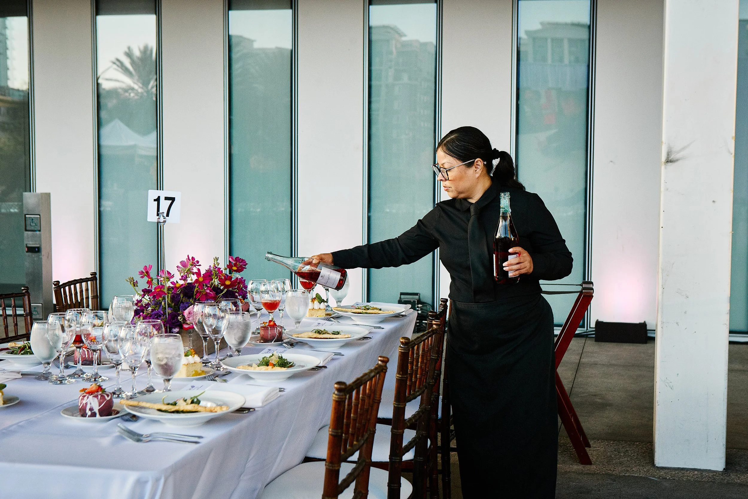 A waitress in a black uniform pouring red wine into glasses at a formal dining table with white tablecloth, flower centerpiece, and multiple glasses and plates, numbered table 17, in an indoor setting with large glass windows.