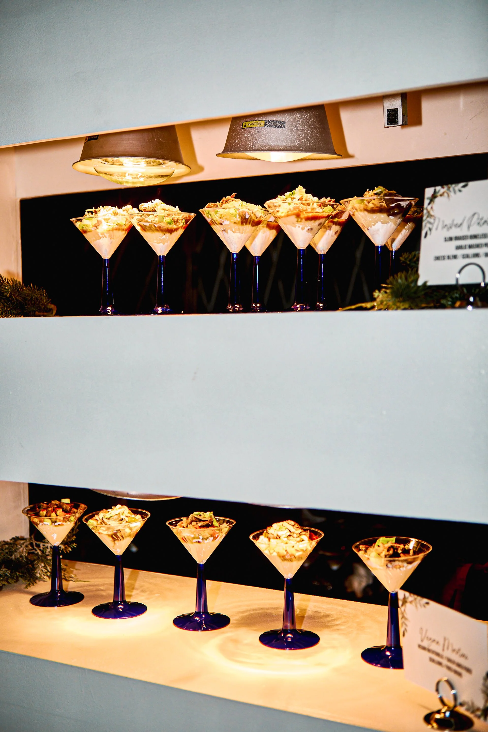Display of multiple dessert parfaits in martini glasses on a two-tiered serving station.