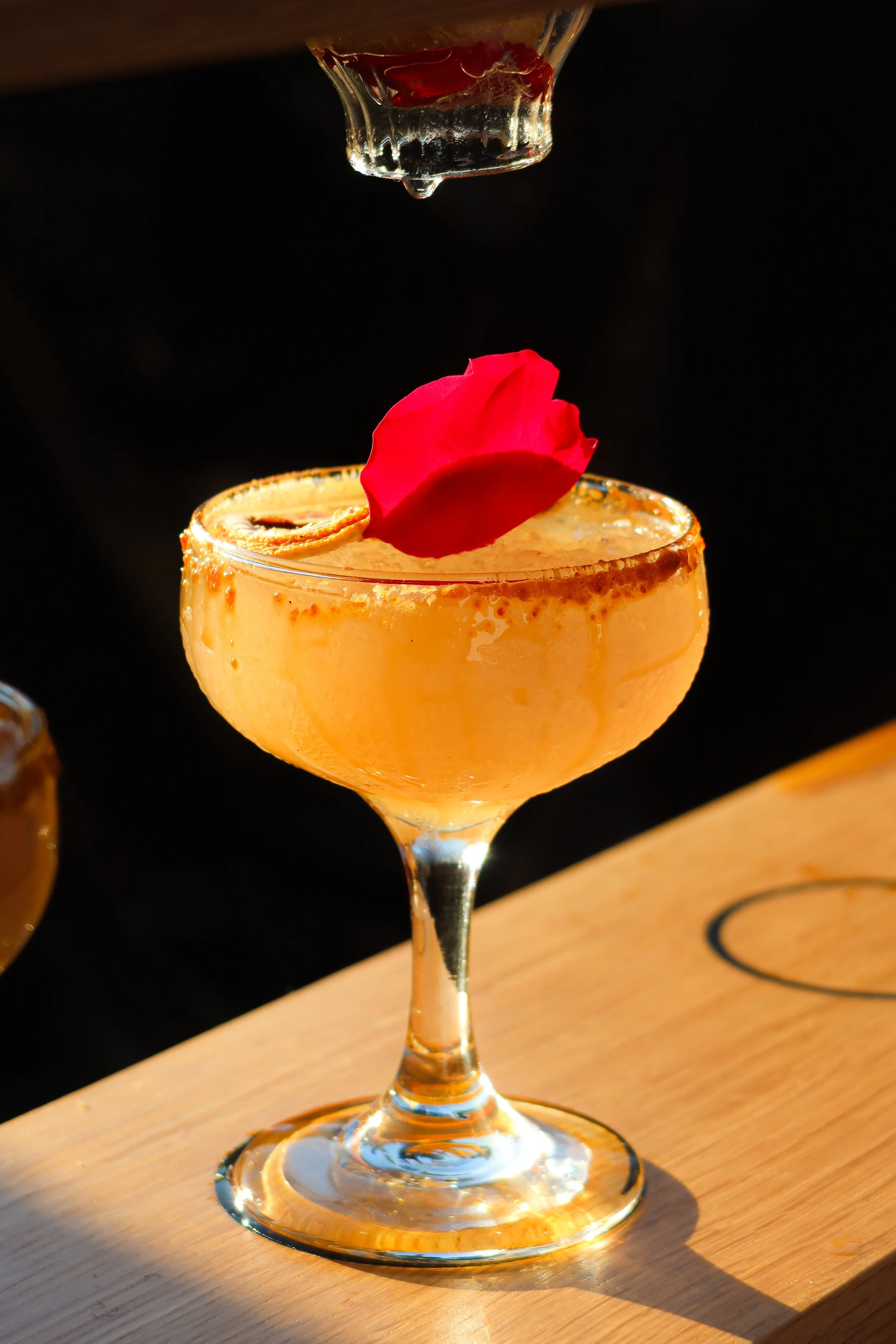 A cocktail in a coupe glass garnished with a red rose petal, with a second glass being poured with liquid above it, on a wooden surface with sunlight and shadows.