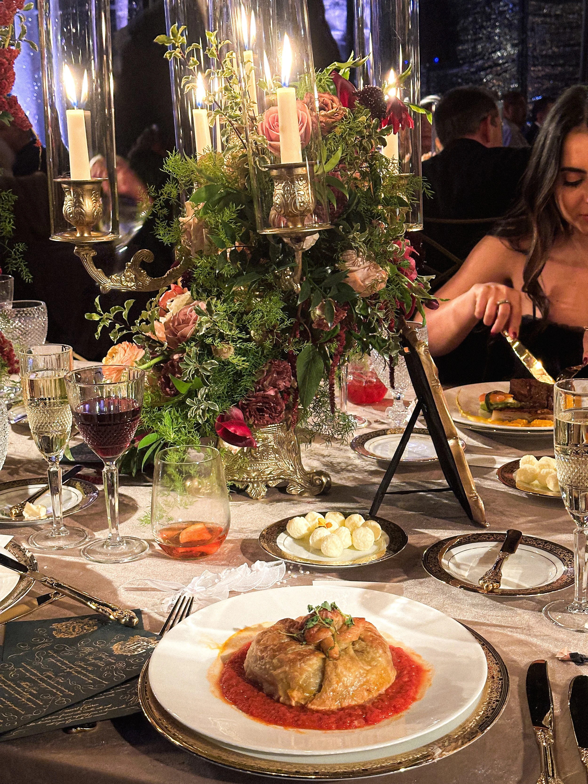 Elegant dinner table with a floral centerpiece, candles, glasses of wine and champagne, a plate of stuffed eggplant, and various other dishes in a dark, upscale setting.