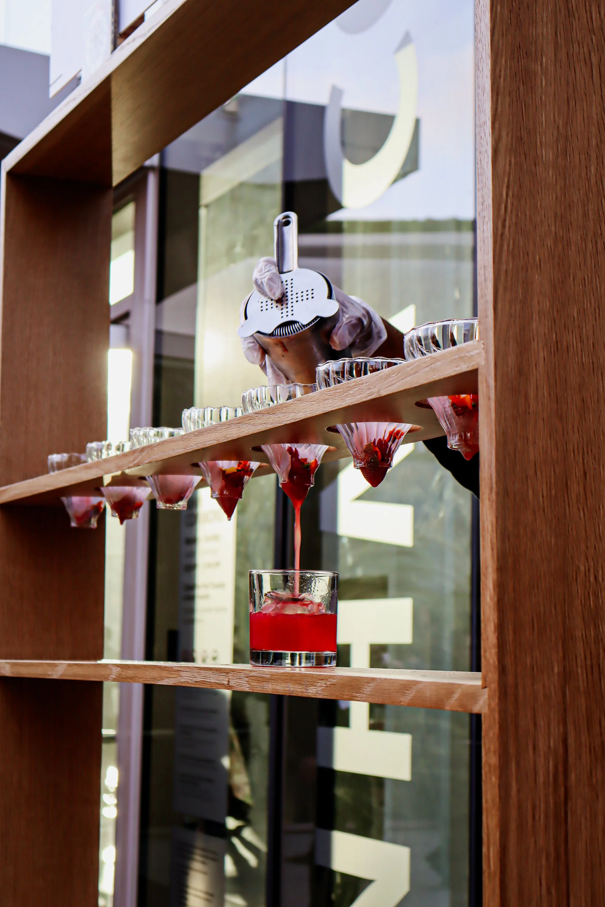 A person is pouring a red liquid from a glass bowl through a strainer into a short glass container, with several small glasses on a wooden shelf behind.
