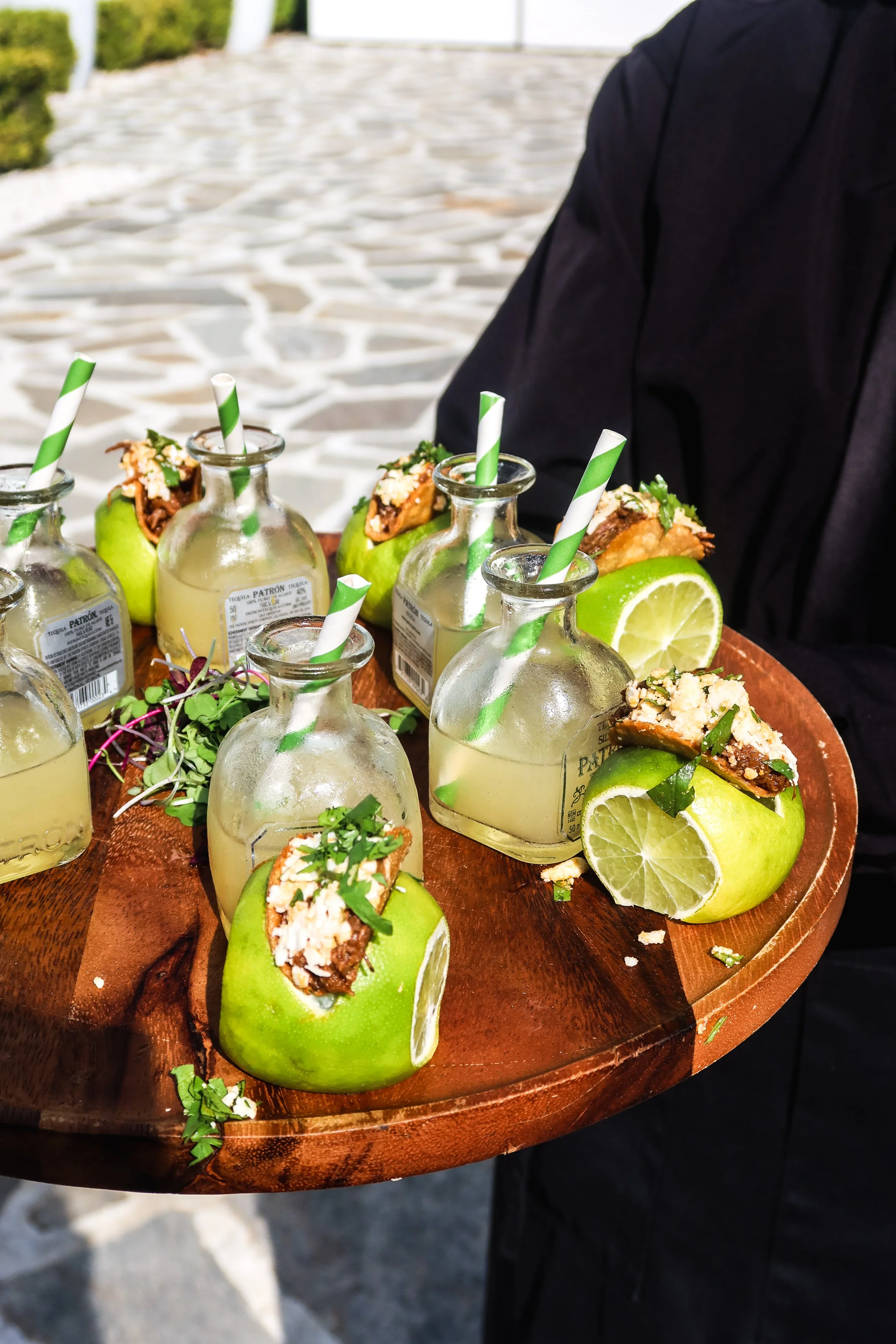 A waiter holds a round wooden tray with shot glasses filled with a light-colored beverage, garnished with cilantro and lime wedges, and stuffed green apples with tacos and green-striped straws.