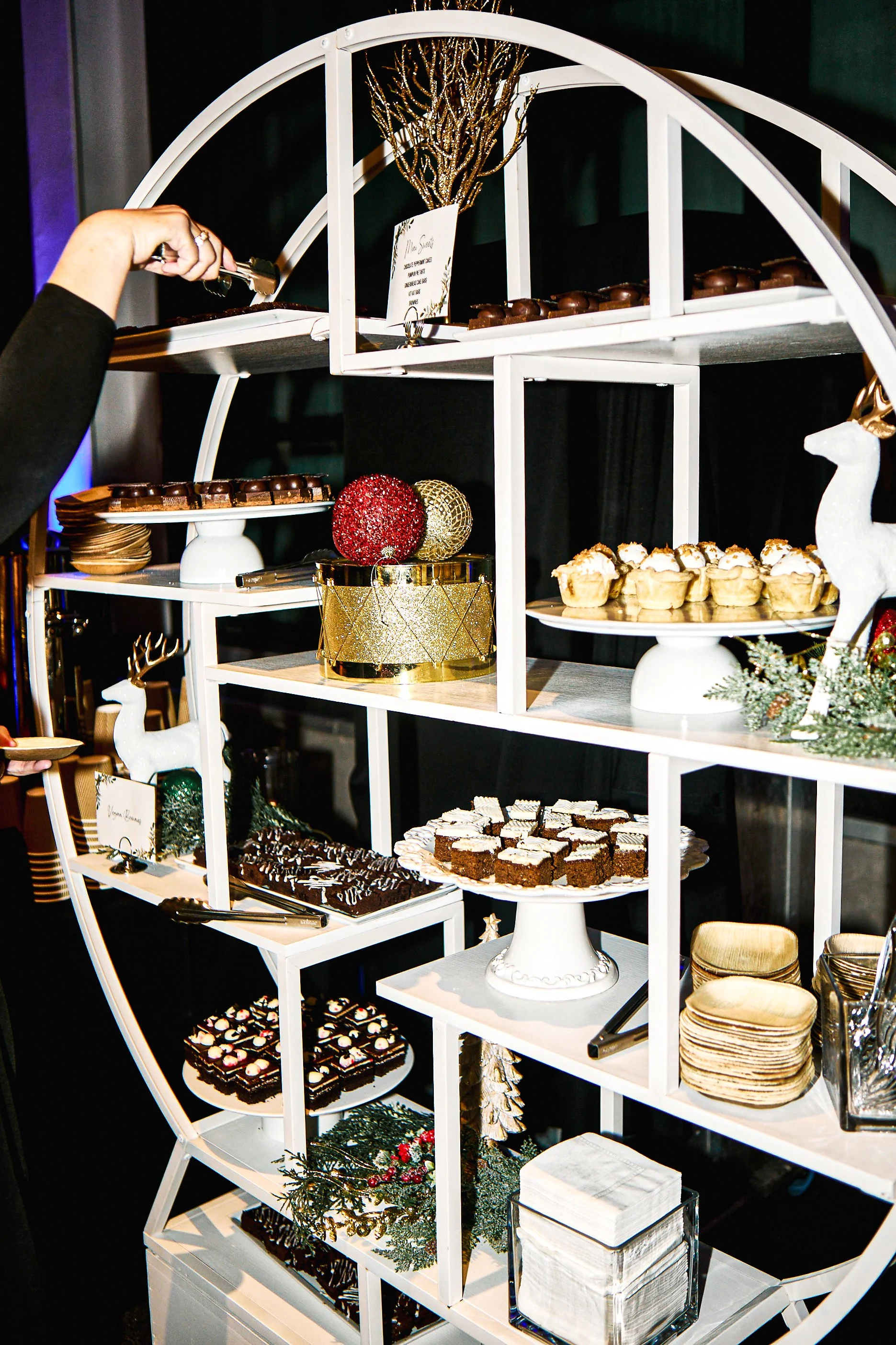 A white carousel-style dessert display stand with various baked goods and decorations, including cupcakes, brownies, chocolates, and holiday ornaments.