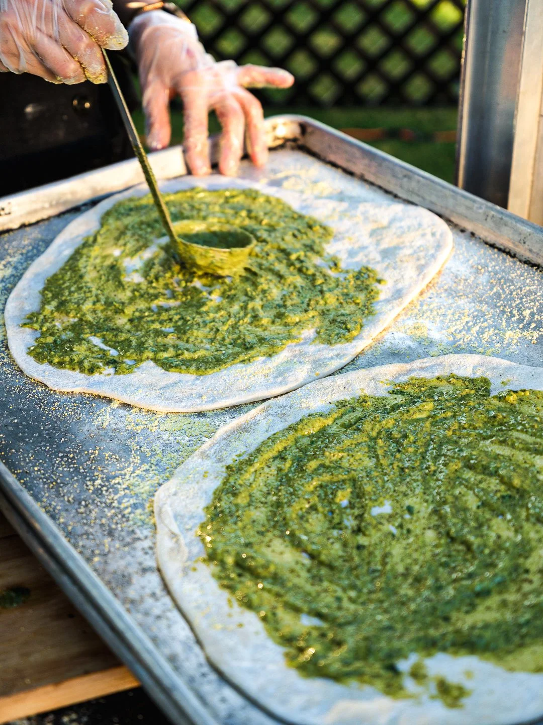 Person preparing pesto on a baking sheet with parchment paper, using a spoon to spread green pesto sauce on flatbread or dough outdoors.