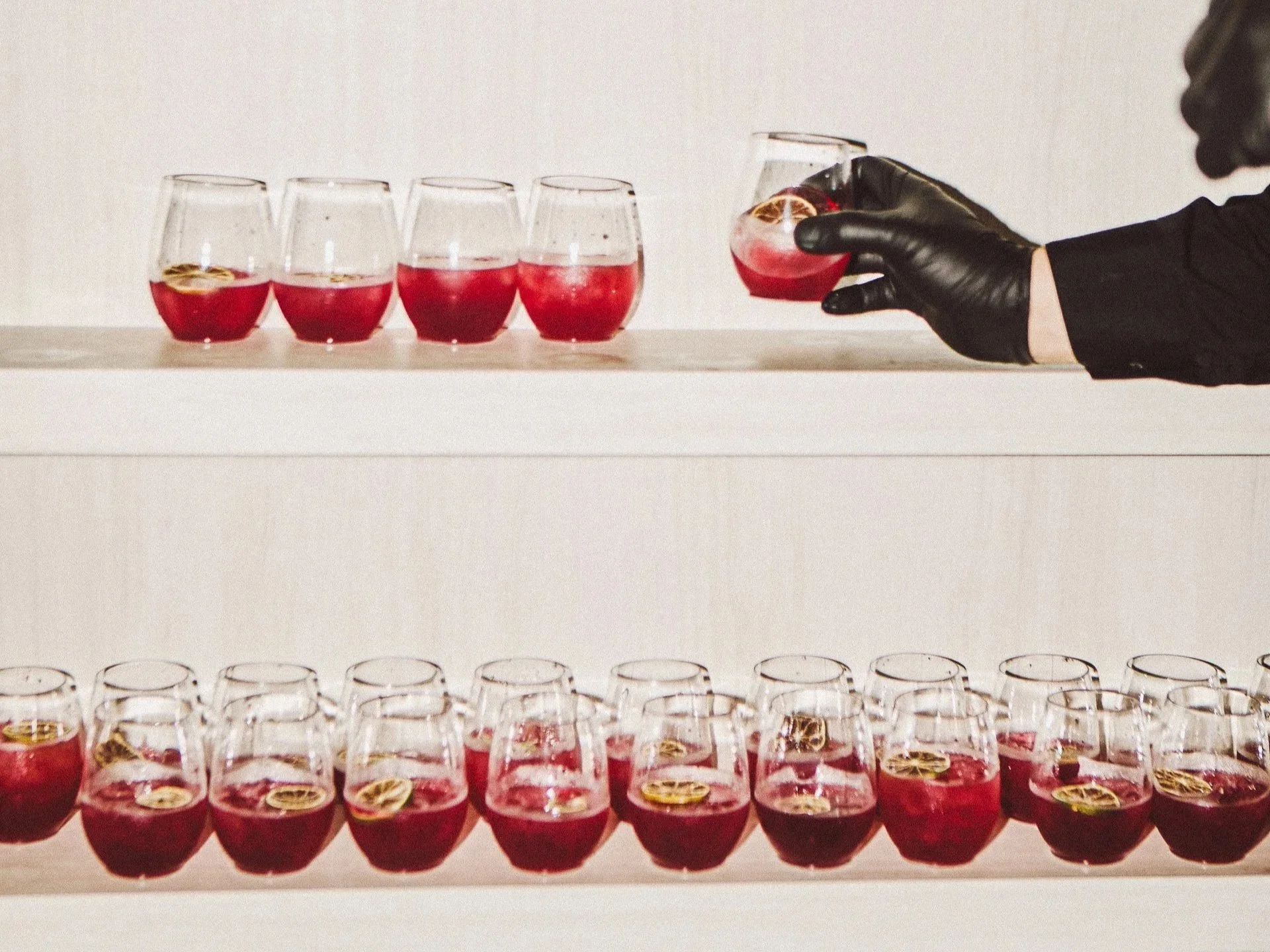 A person wearing black gloves and a black shirt holding a glass of red cocktail with lemon slices, with multiple glasses of similar drinks on two white shelves, some with lemon slices inside.