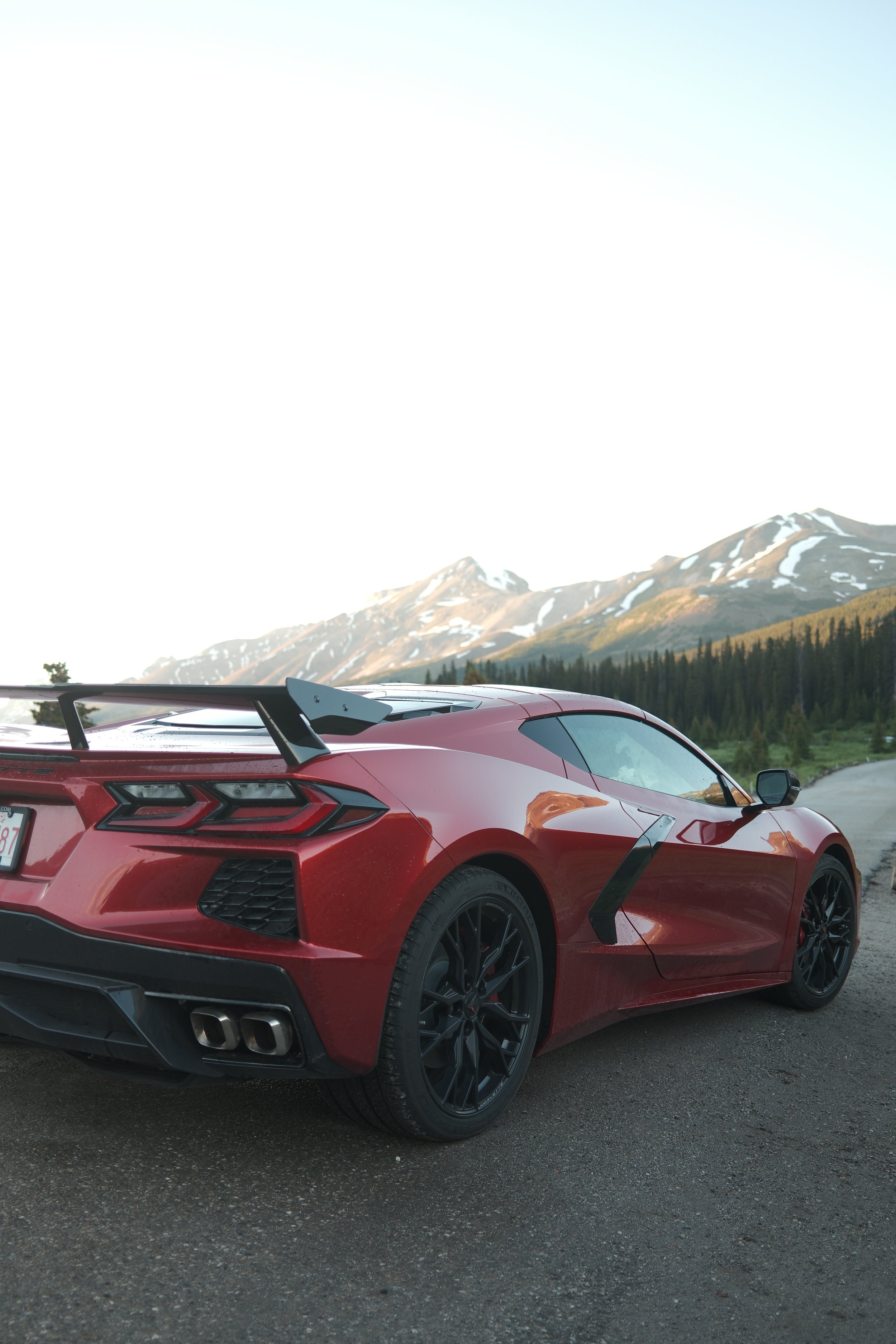 A red sports car with black wheels parked on a mountain road. The car has a spoiler and dual exhaust pipes. In the background, there are mountains with patches of snow and a forest of trees.