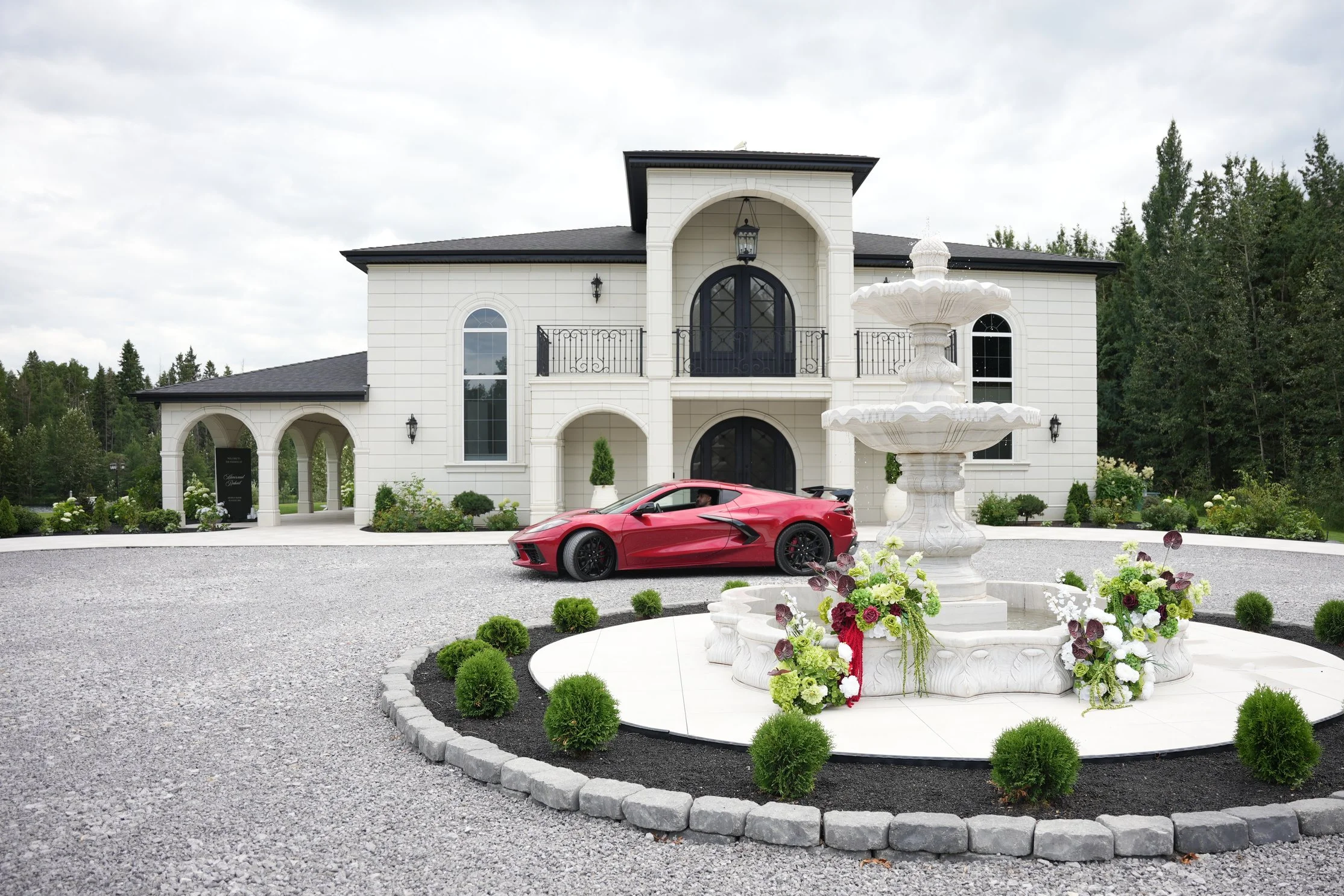 A red sports car parked in front of a white mansion with black accents, surrounded by a gravel driveway, a fountain with flowers, and lush green trees in the background.