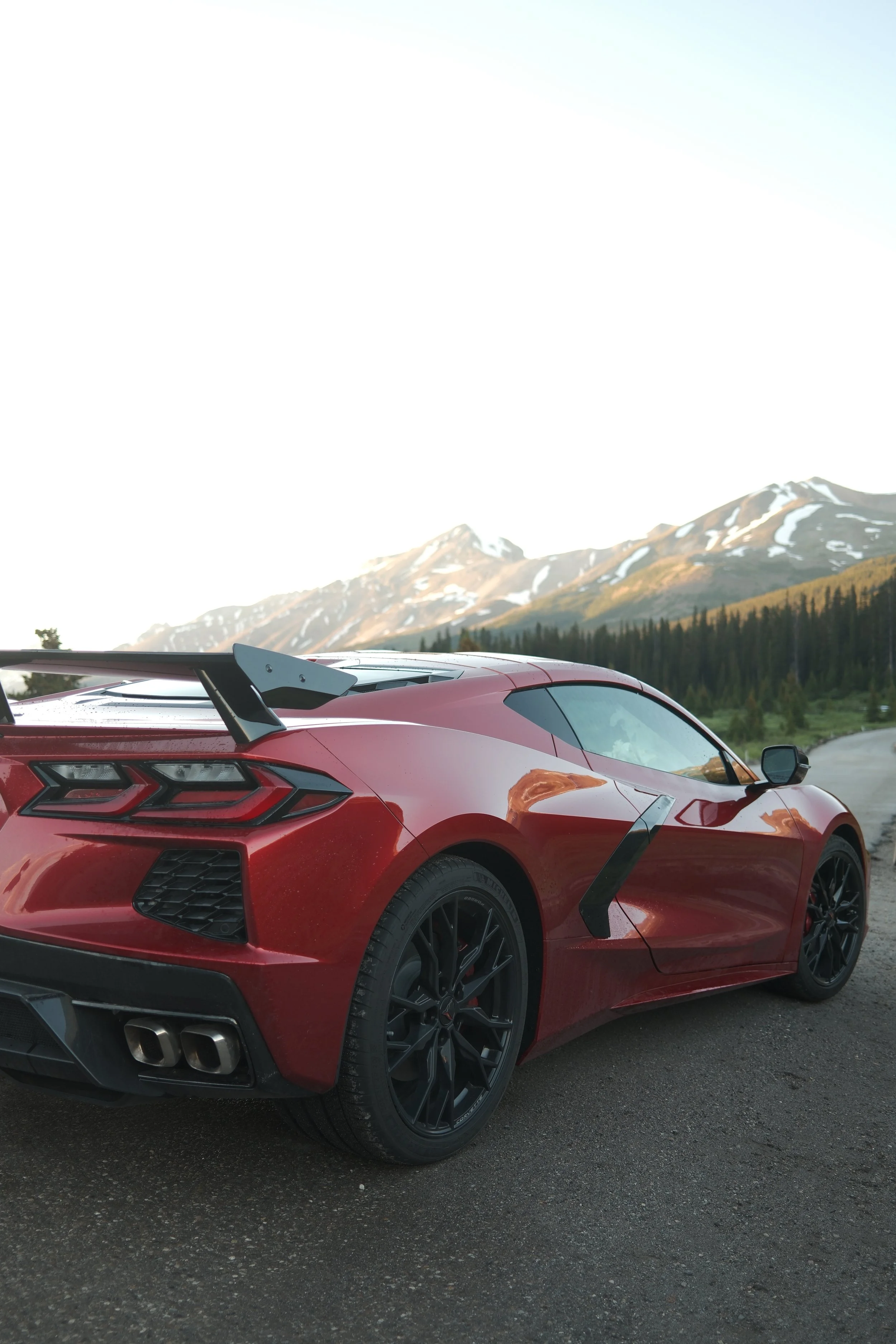 Red sports car with black wheels and rear spoiler parked on the side of a road against a backdrop of snow-capped mountains and a forest.