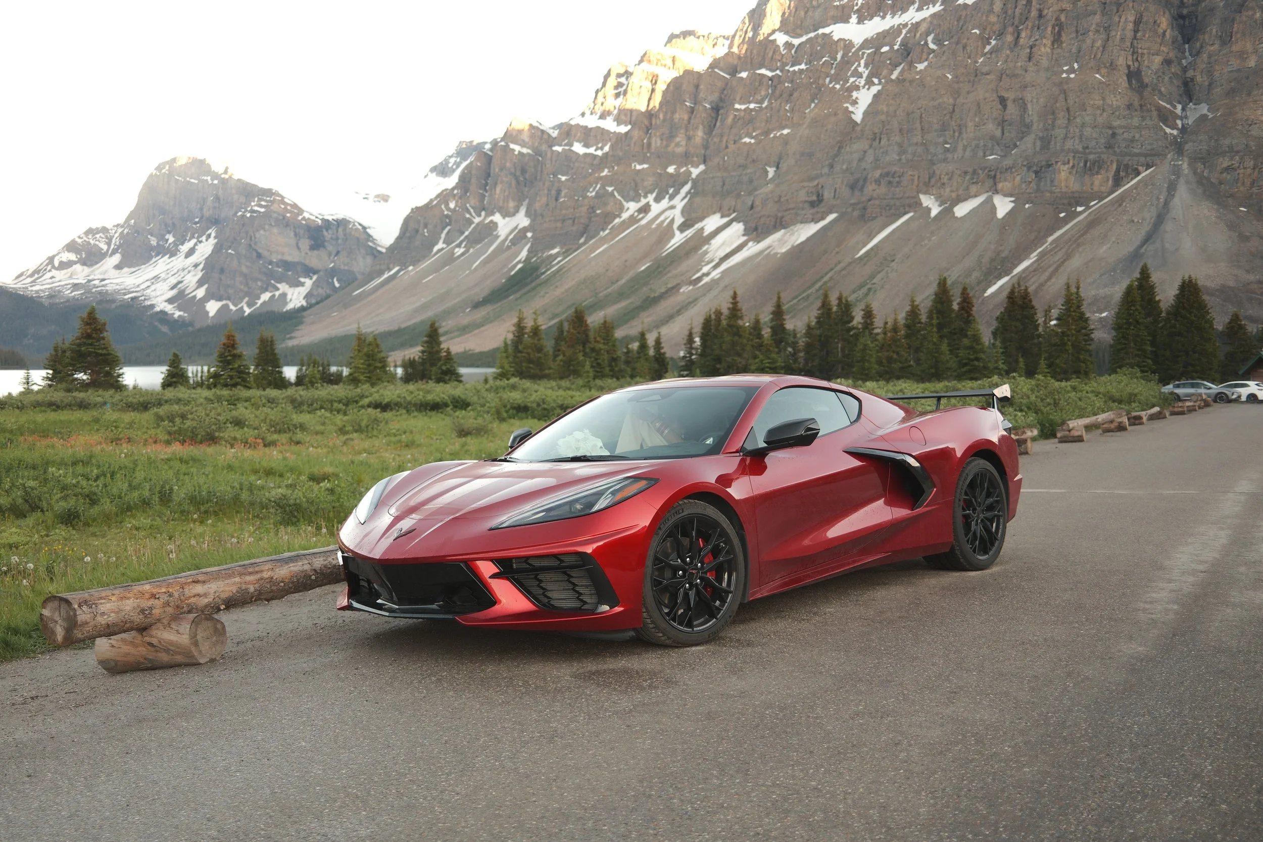 Red sports car parked outdoors near logs with a background of mountains, trees, and a lake.