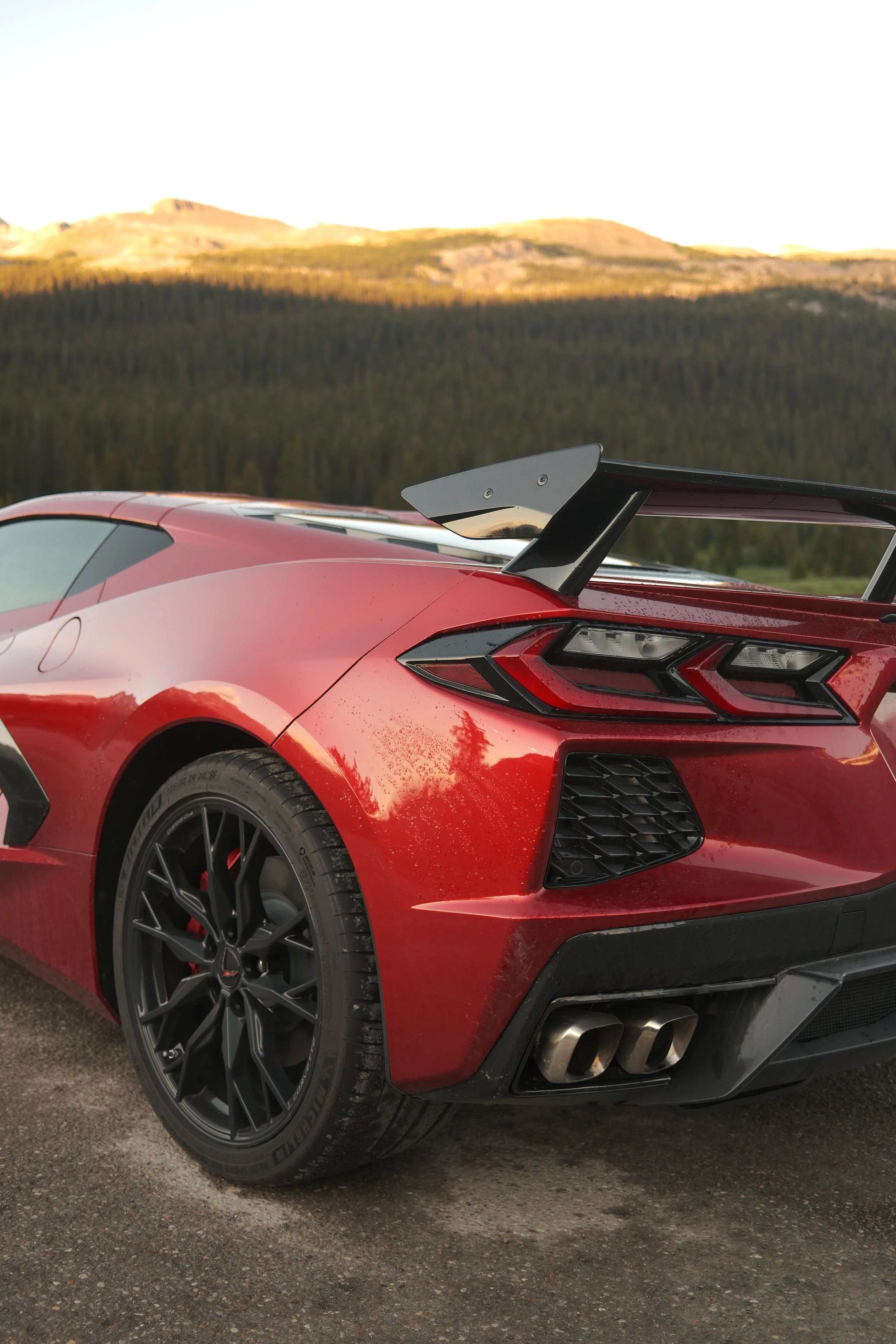 A red sports car with a black rear spoiler and black wheels parked on a gravel road, with a forest and mountains in the background during sunset.