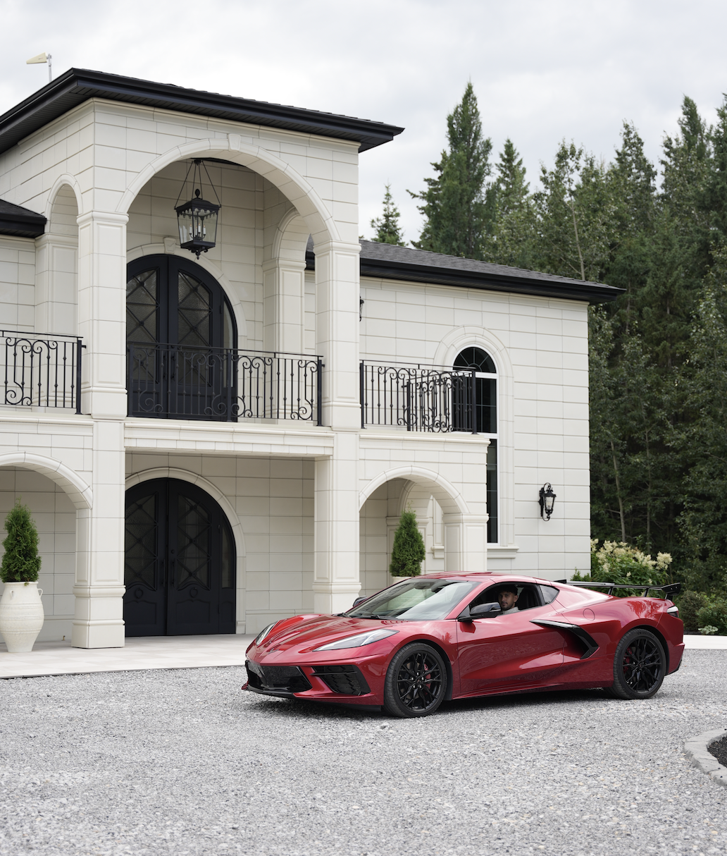 A red sports car parked in front of a large, white, two-story house with a black gate and trees in the background.