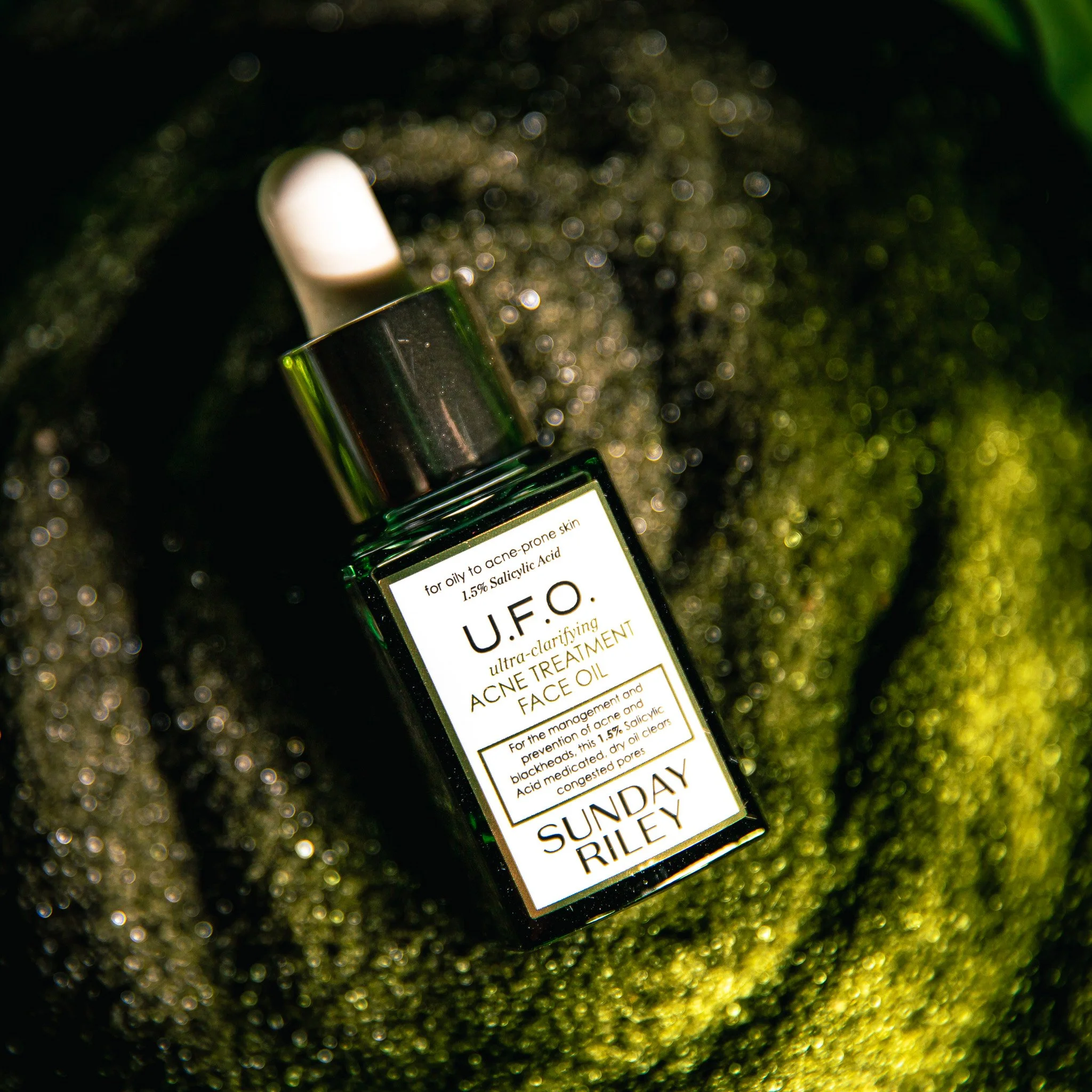 A small, dark green glass bottle with a white dropper top and a label that reads 'Sunday Riley U.F.O.' sitting in dark, shimmering black sand with green light leaking onto it.