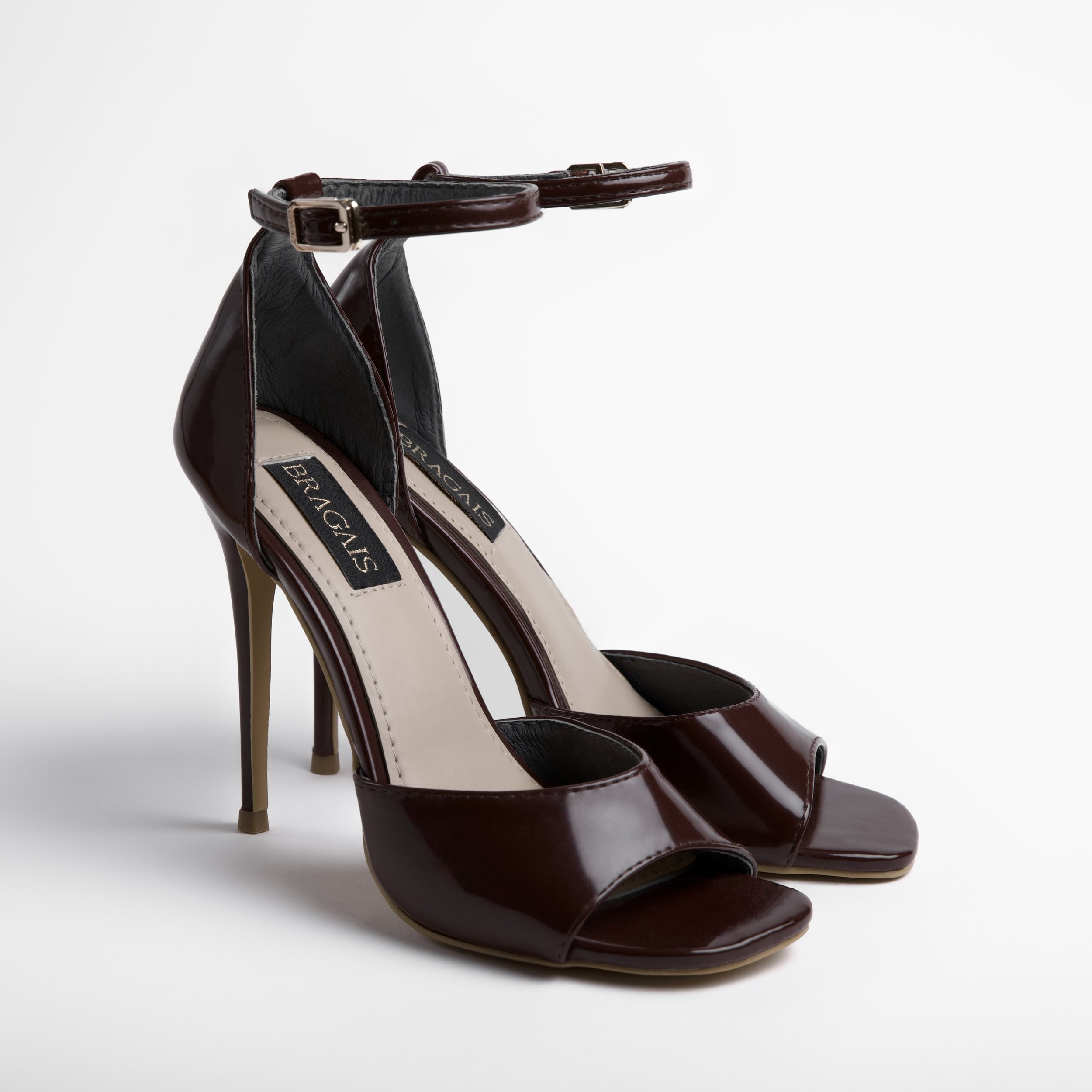 Close-up of a pair of high-heeled women's stiletto pageant shoes with dark maroon patent leather and ankle straps, placed on a white surface.