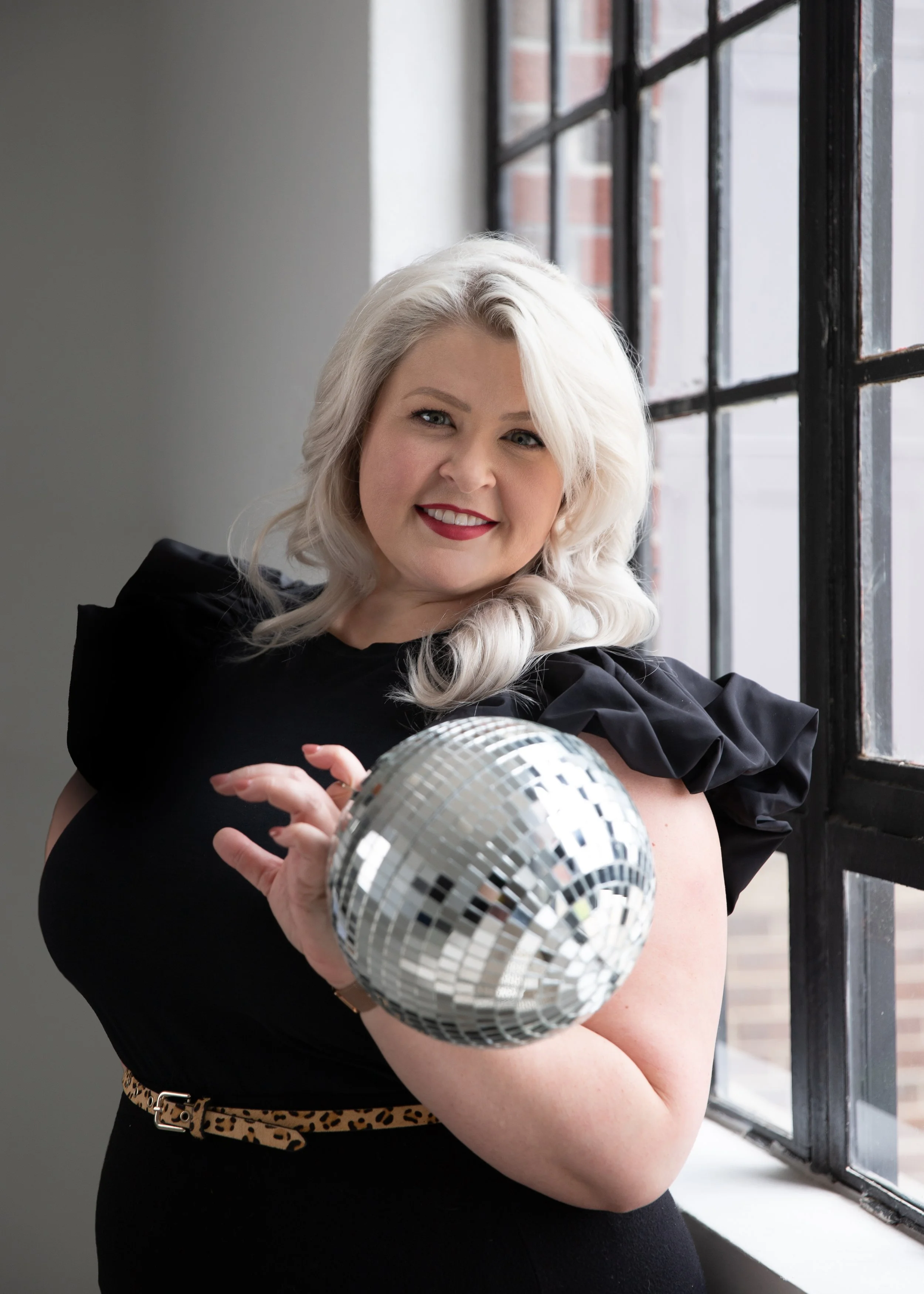 A woman with blonde hair, wearing a black dress and leopard print belt, holding a disco ball, standing by a window with black metal framing and brick wall outside.