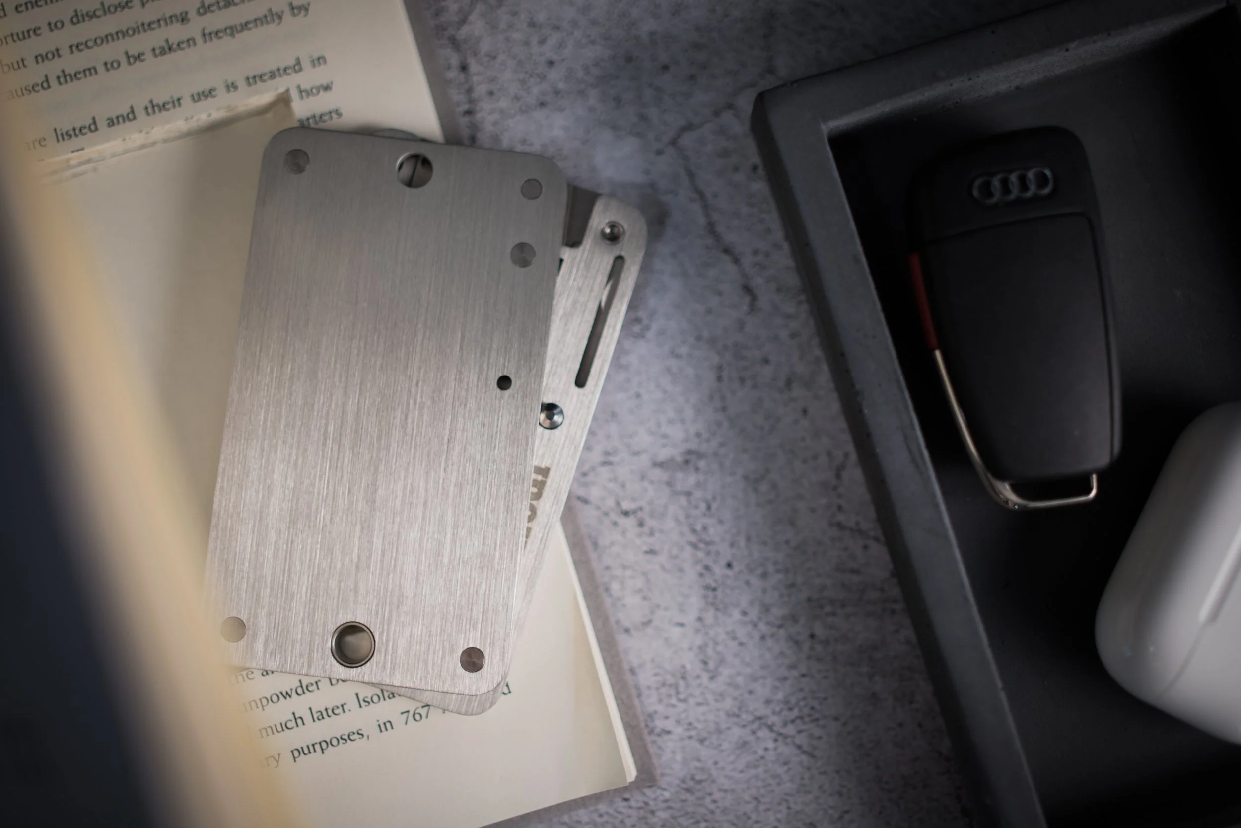 A metal plate with multiple holes and a slot resting on a book page with printed text, next to a black Audi car key.