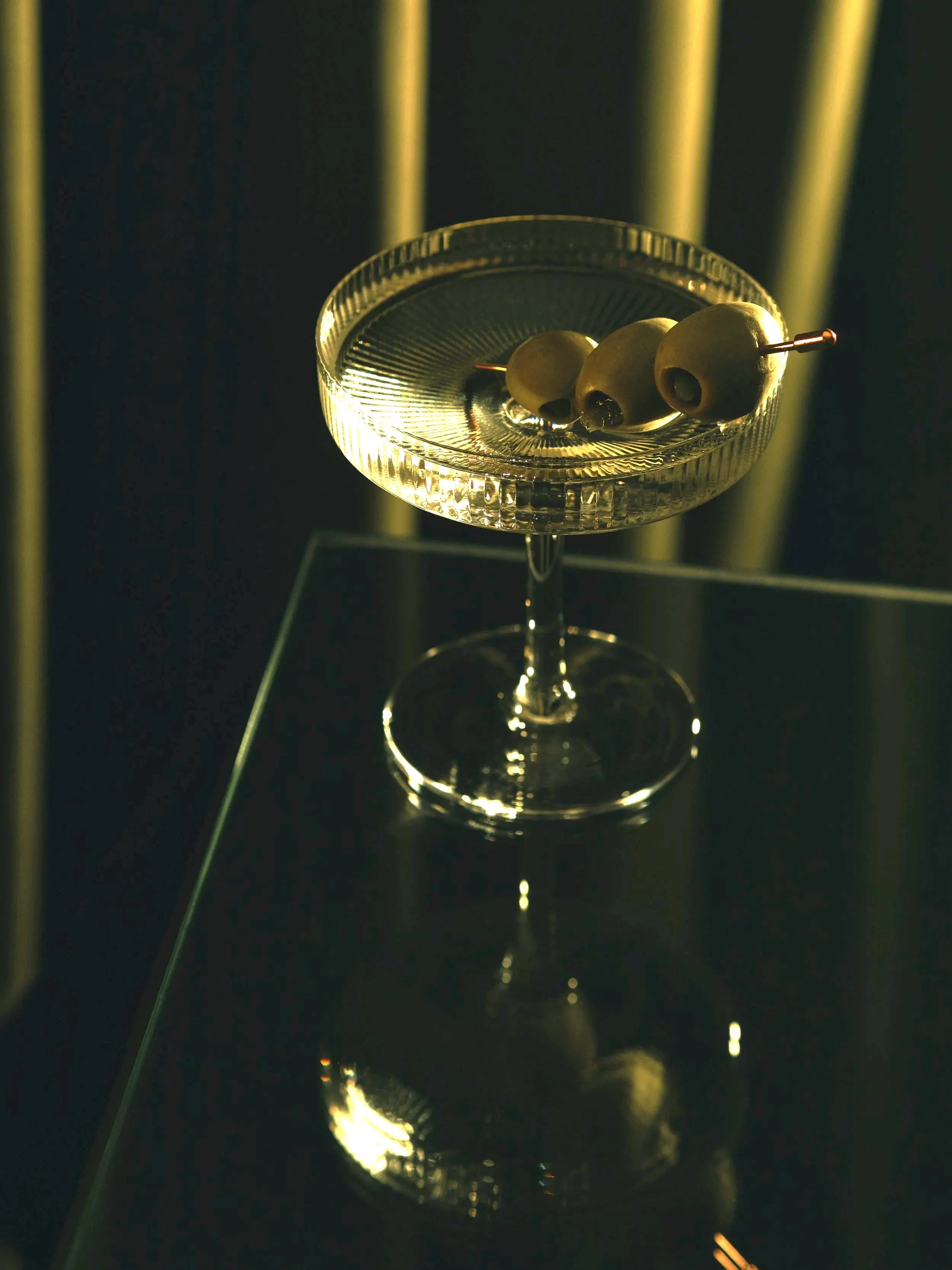 Three green olives on a metal cocktail pick resting on the glass rim of a martini that's sitting on a reflective black surface with a dark green curtain background.