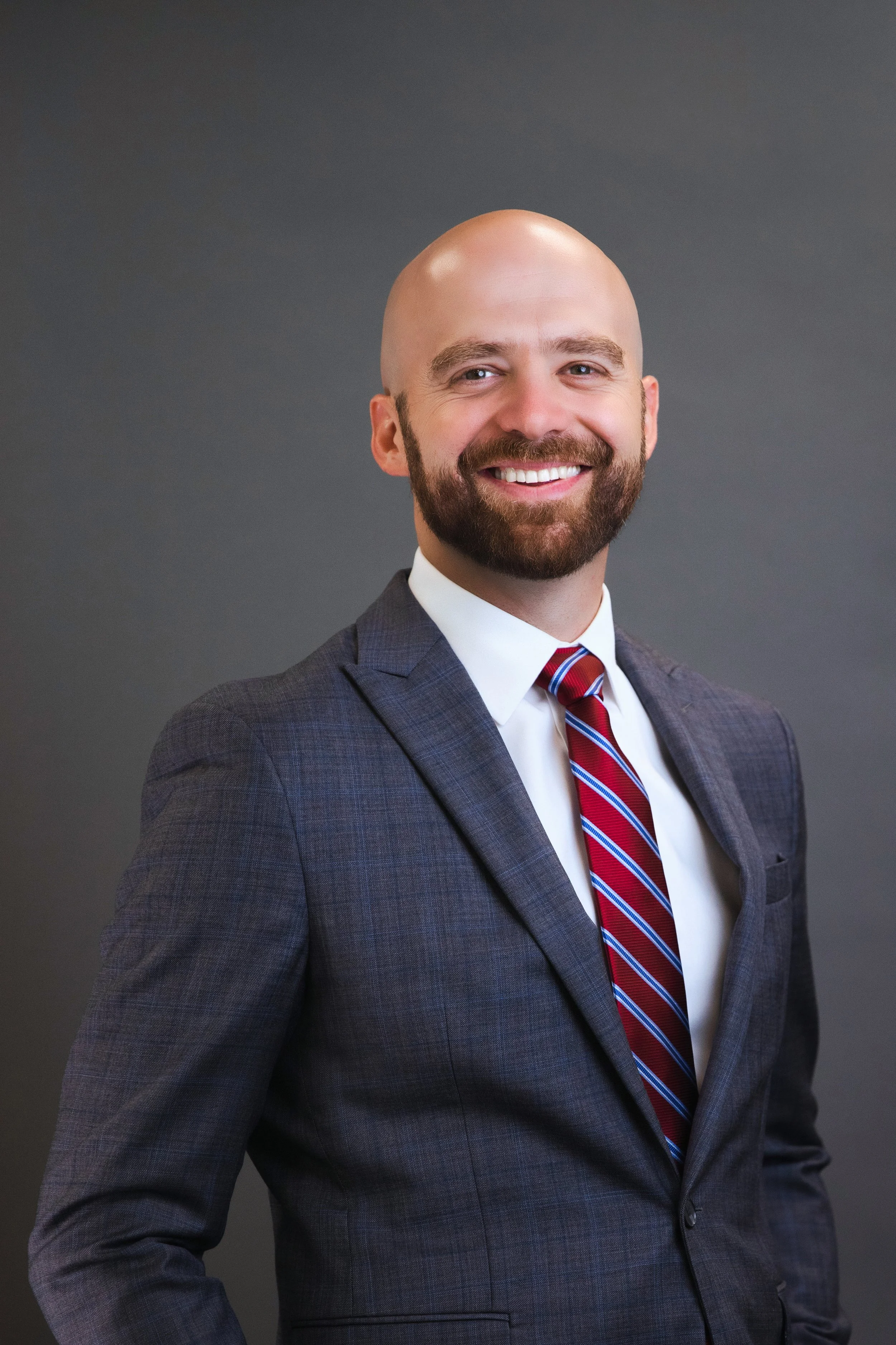 A man in a gray suit, white shirt, and red striped tie smiling against a gray background.