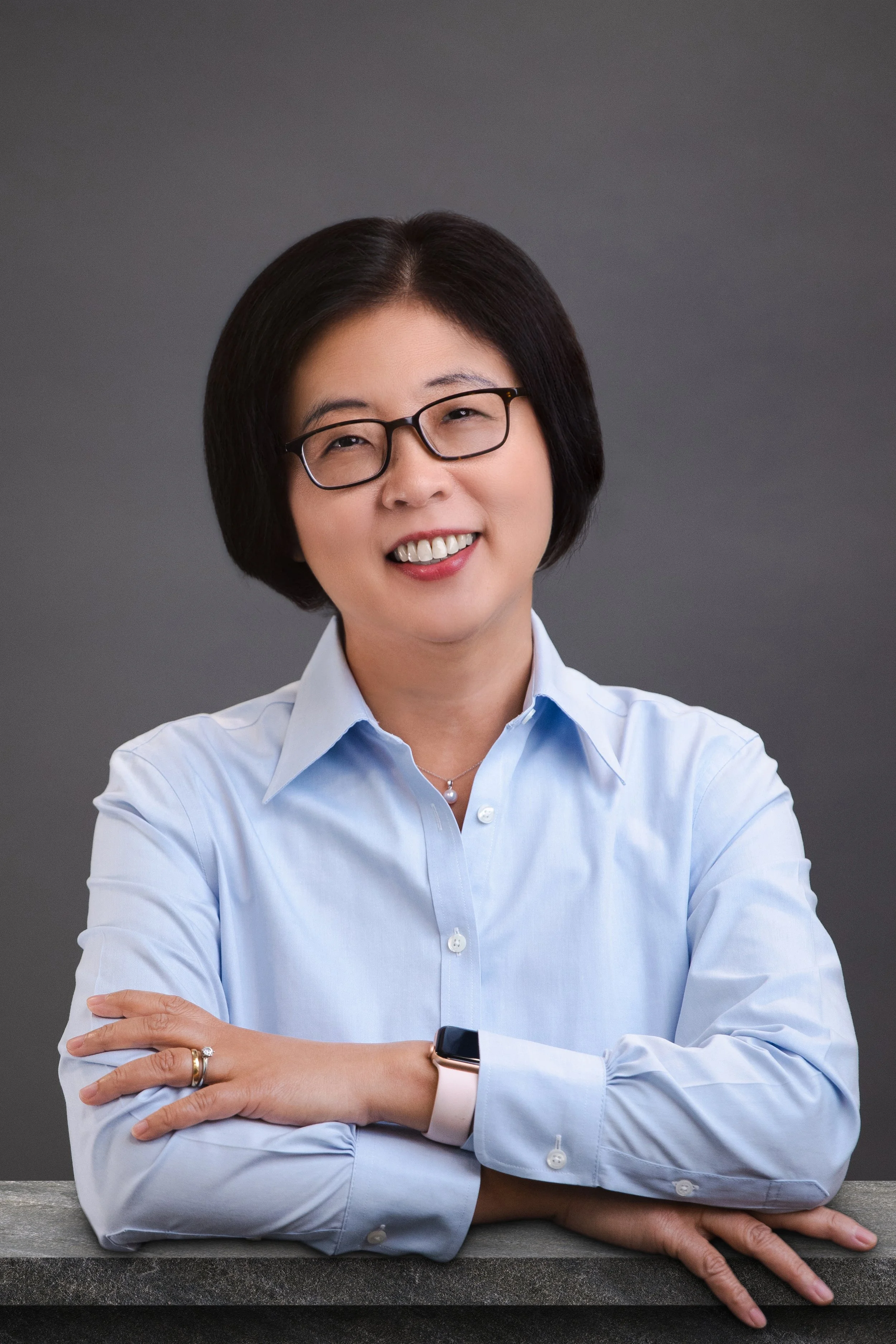 A woman with short dark hair and glasses wearing a light blue button-up shirt. She is smiling, crossing her arms, and wearing a watch on her left wrist. She is sitting at a gray textured surface with a gray gradient background.