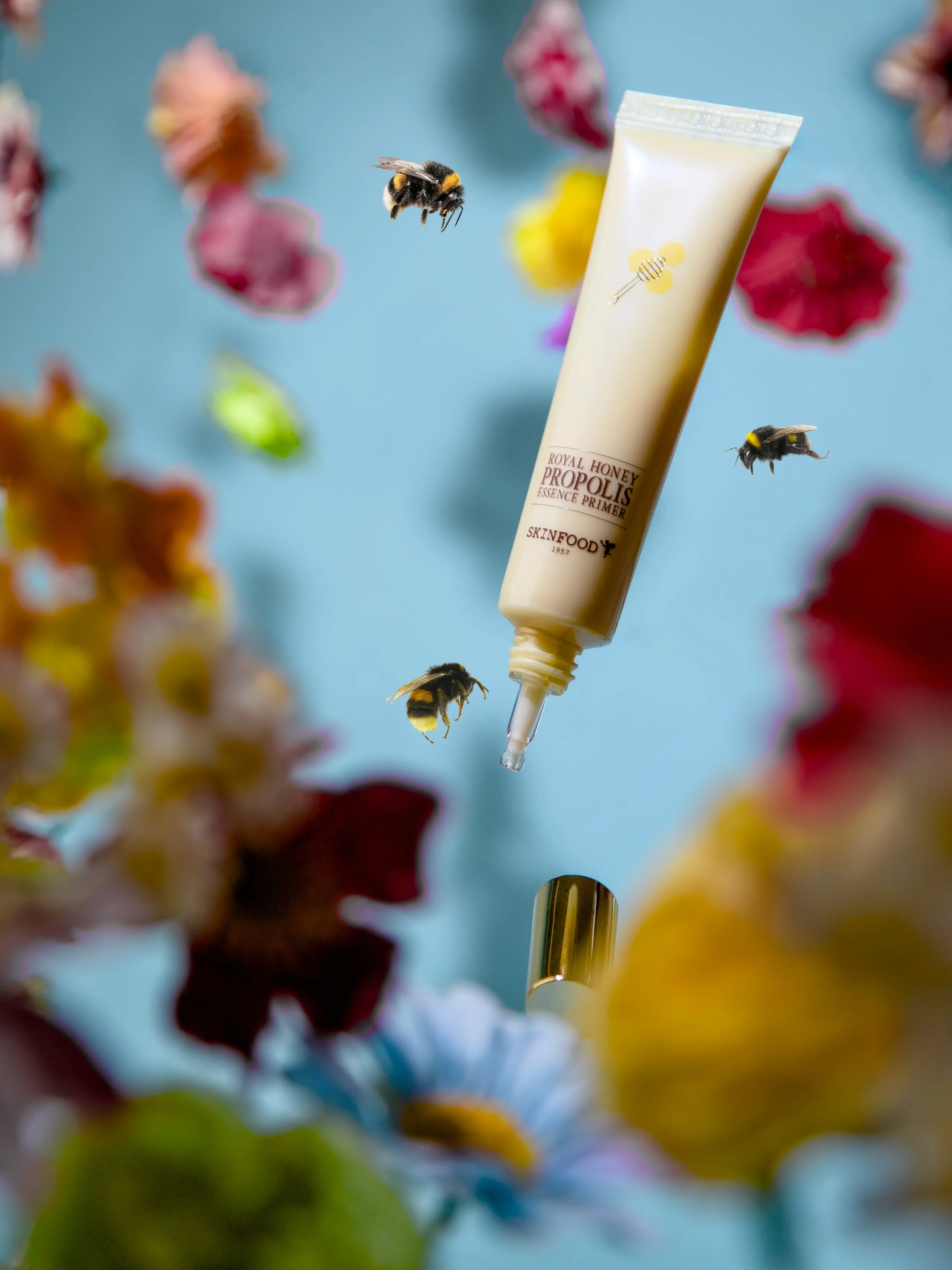 A tube of Skinfood Royal Honey Propolis Essence Primer surrounded by blurred colorful flowers and bees in flight against a blue background.