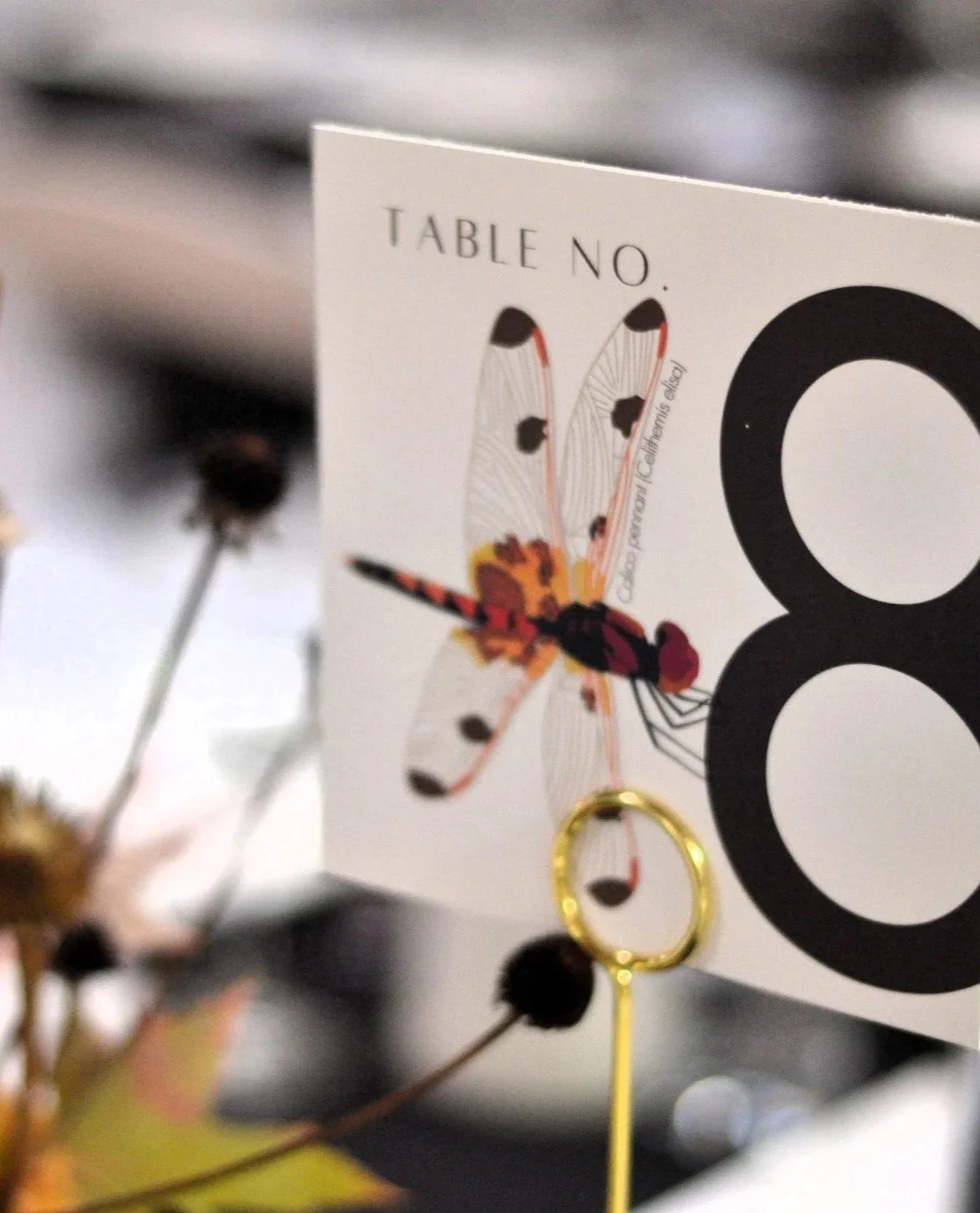 A close-up of a table number card with a colorful dragonfly illustration perched on the number "8" and the text 'TABLE NO.' at the top, surrounded by blurred flowers and decorative table elements.