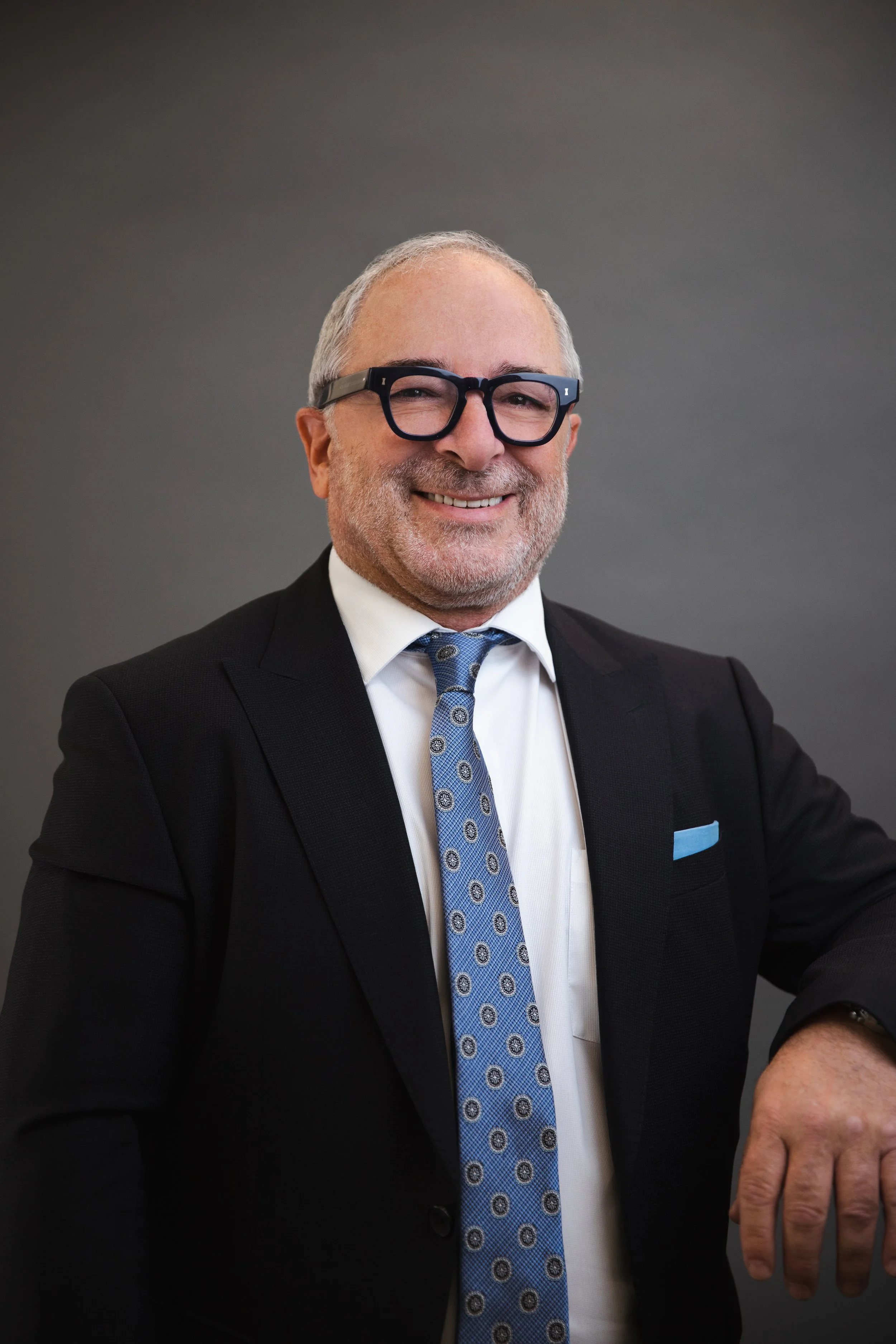 A smiling man with gray hair, beard, and glasses wearing a black suit, white shirt, and blue patterned tie, against a plain gray background.