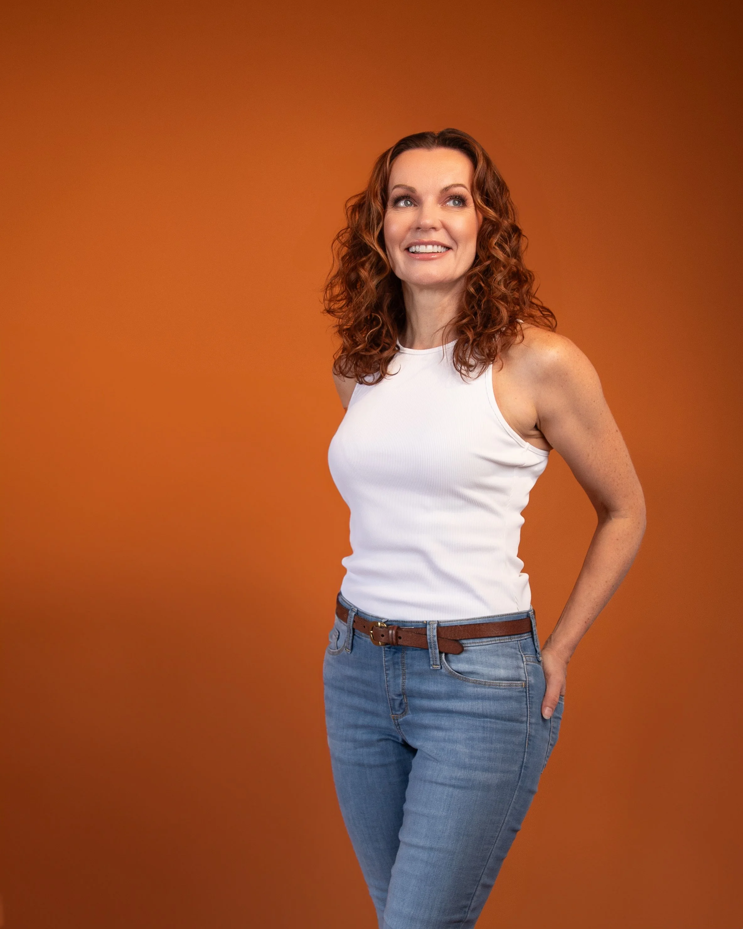 A woman with curly red hair, wearing a white tank top and blue jeans, is standing against a solid orange background, smiling and looking slightly upward.