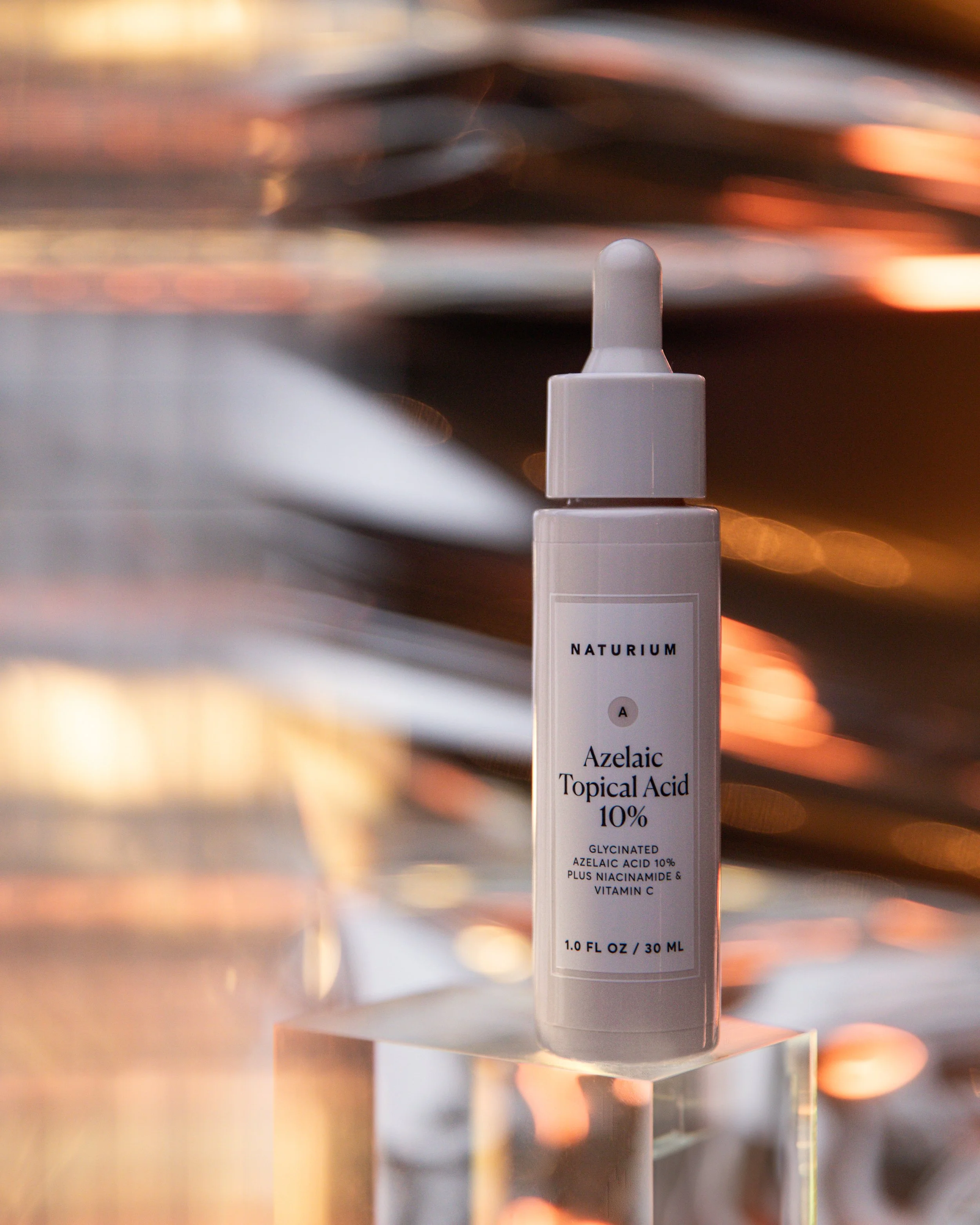 Close-up of a white bottle labeled 'Azelaic Topical Acid 10%', with a white cap, standing on a reflective metallic surface with a blurred background of shiny, warm-colored lights.