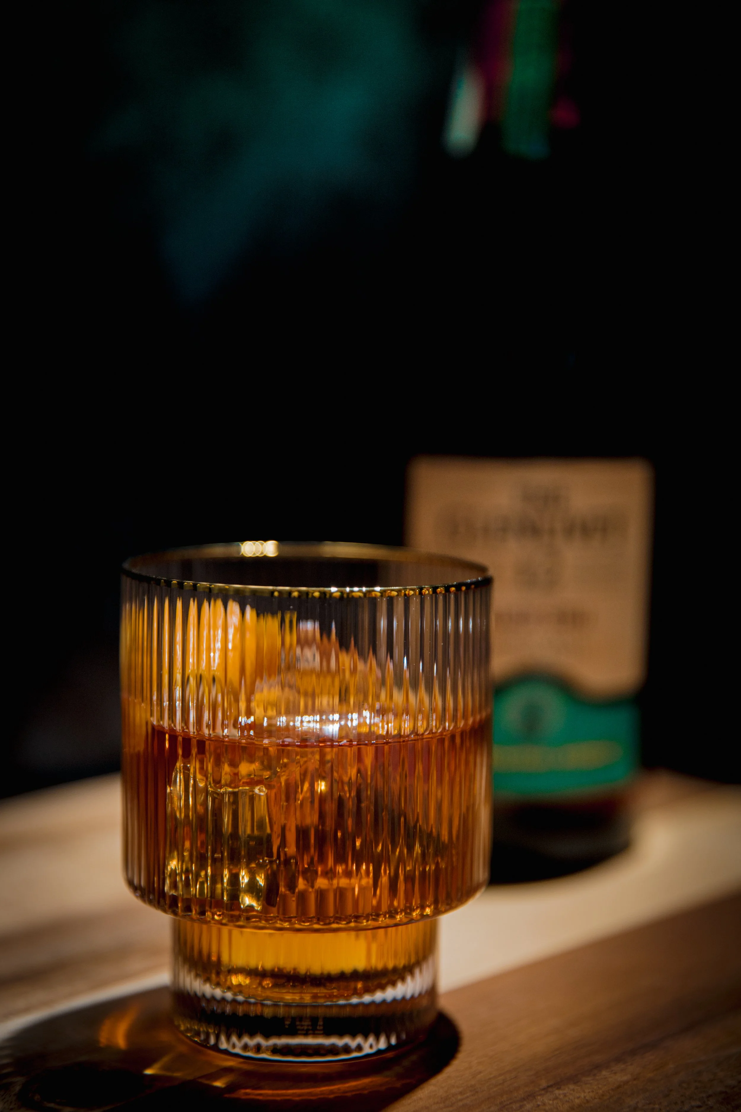 A close-up of a textured amber-colored glass on a wooden surface with a blurred background that includes a green and black object, possibly a product package, and a dark setting with focused lighting on the glass.