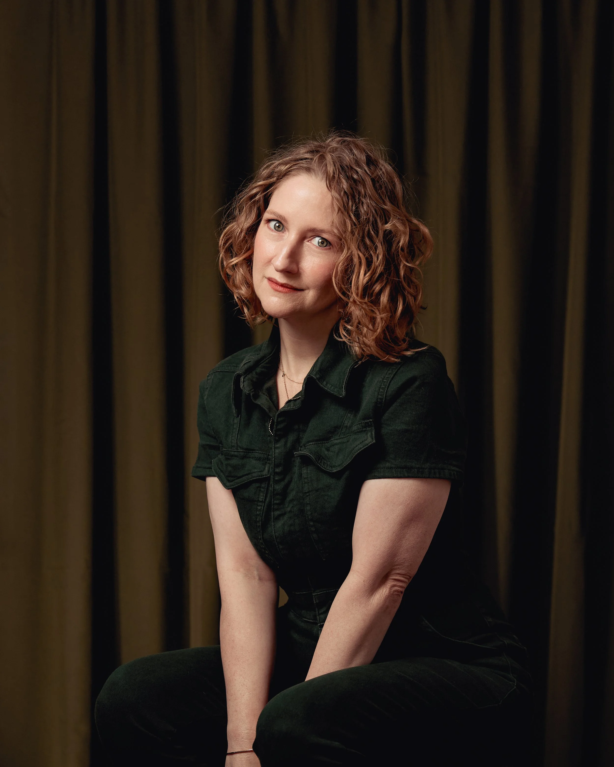 Woman with curly red hair wearing a green denim jumper sitting against a dark curtain background.