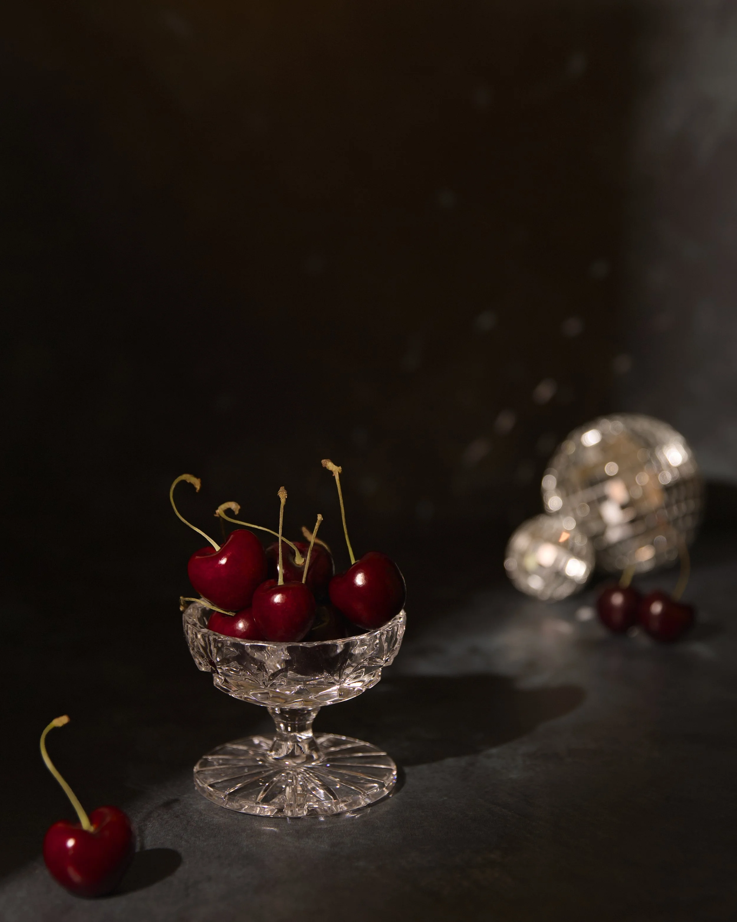 A crystal glass bowl filled with red cherries with stems, placed on a dark surface, with more cherries and blurred, sparkling mini disco balls in the background.