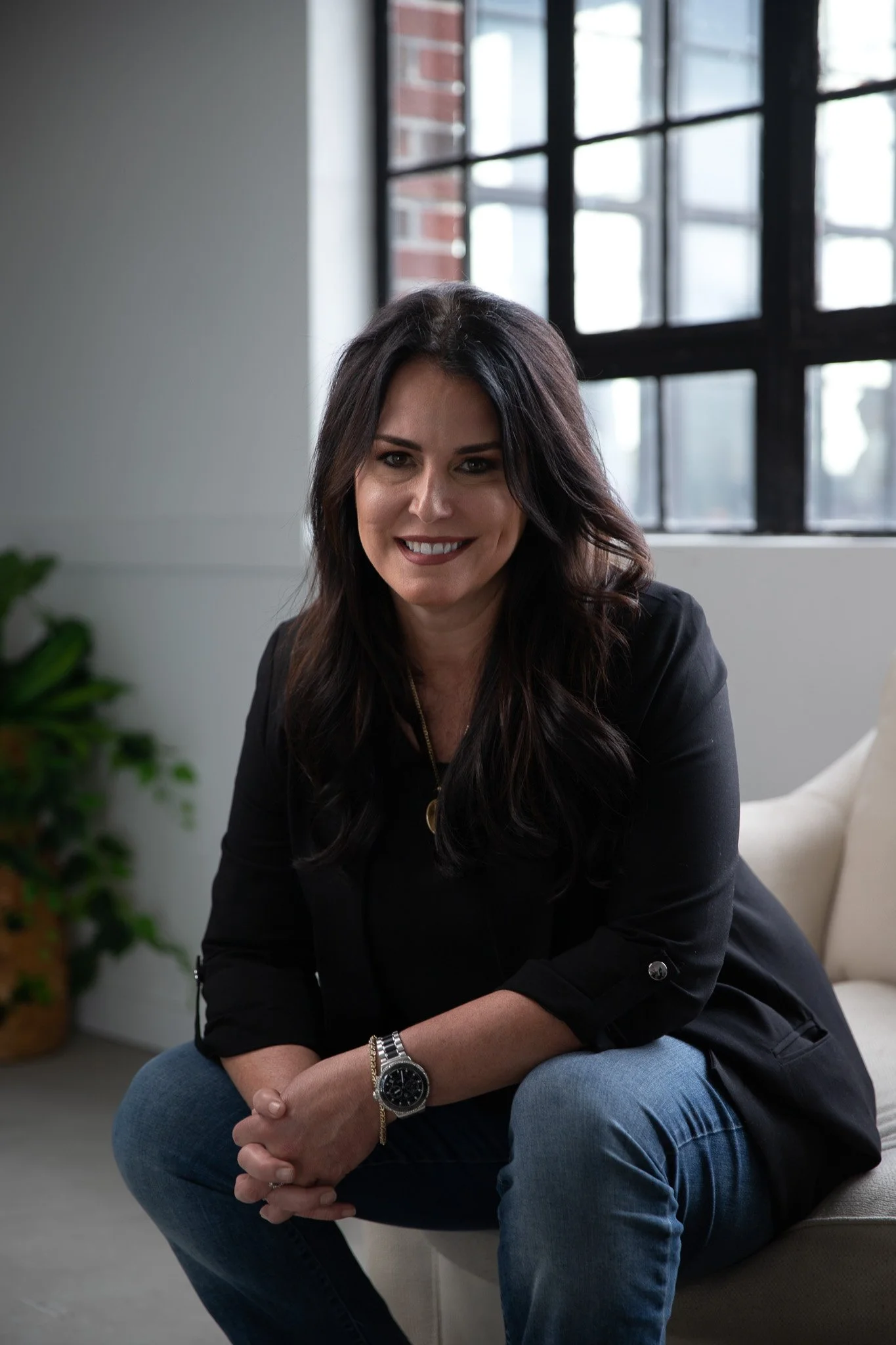 A woman with long dark hair wearing a black jacket and blue jeans, sitting on a beige chair by a large window in a modern room, smiling at the camera.