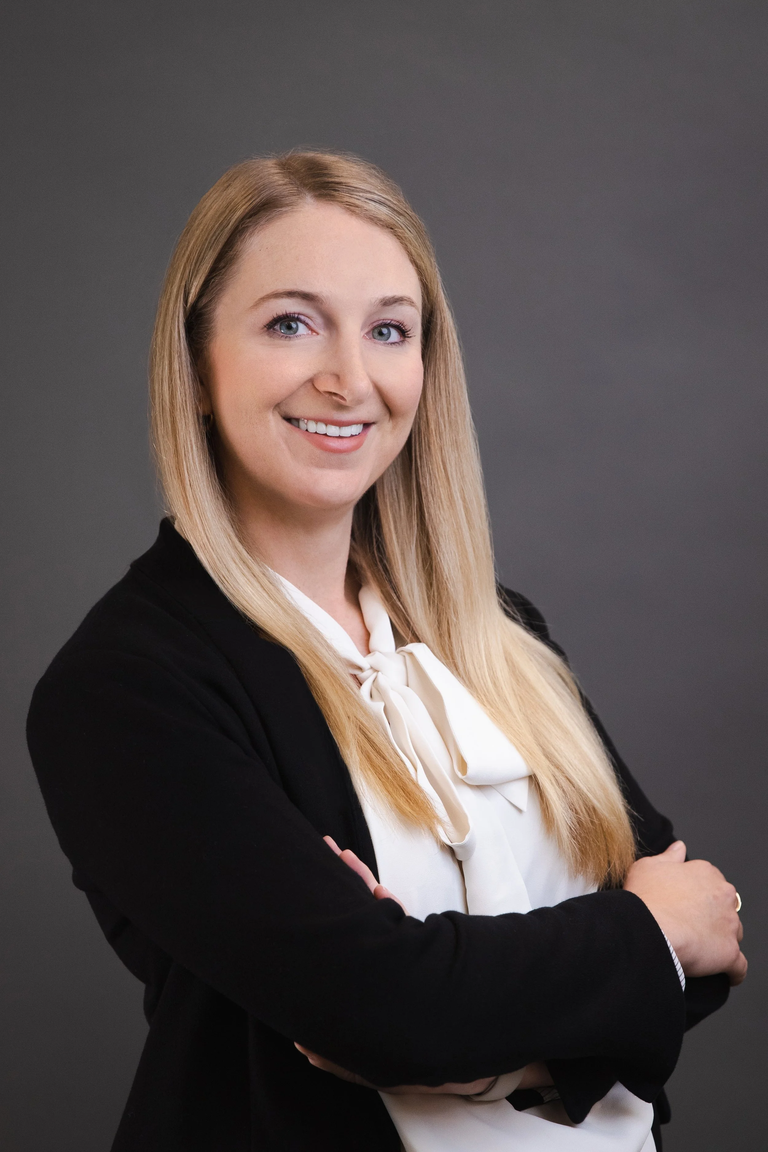 A professional woman with long blonde hair wearing a black blazer and white blouse, smiling with arms crossed against a gray background.
