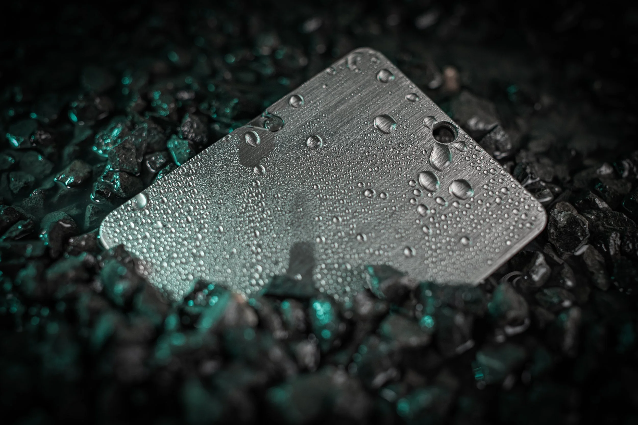 A close-up of a metal card with water droplets on its surface, lying on black gravel rocks.