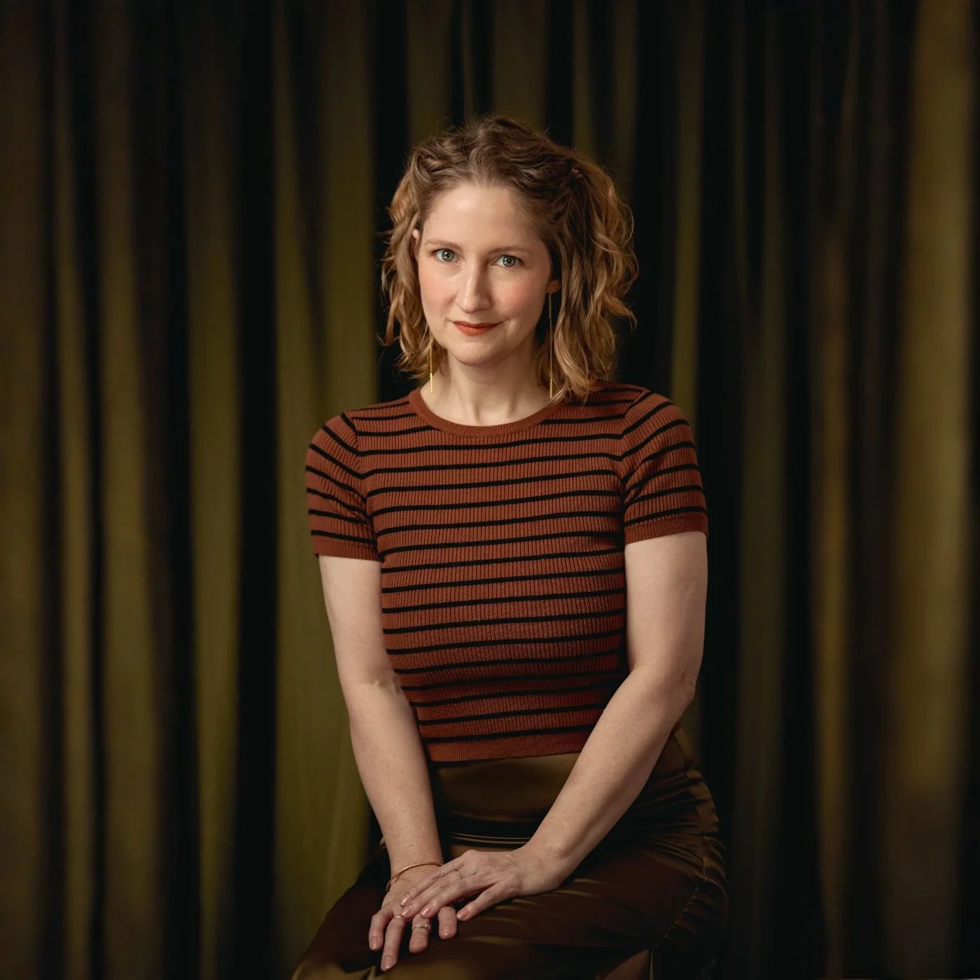 A woman with wavy blonde hair, wearing a rust and black striped shirt, sitting in front of a dark background of curtains.