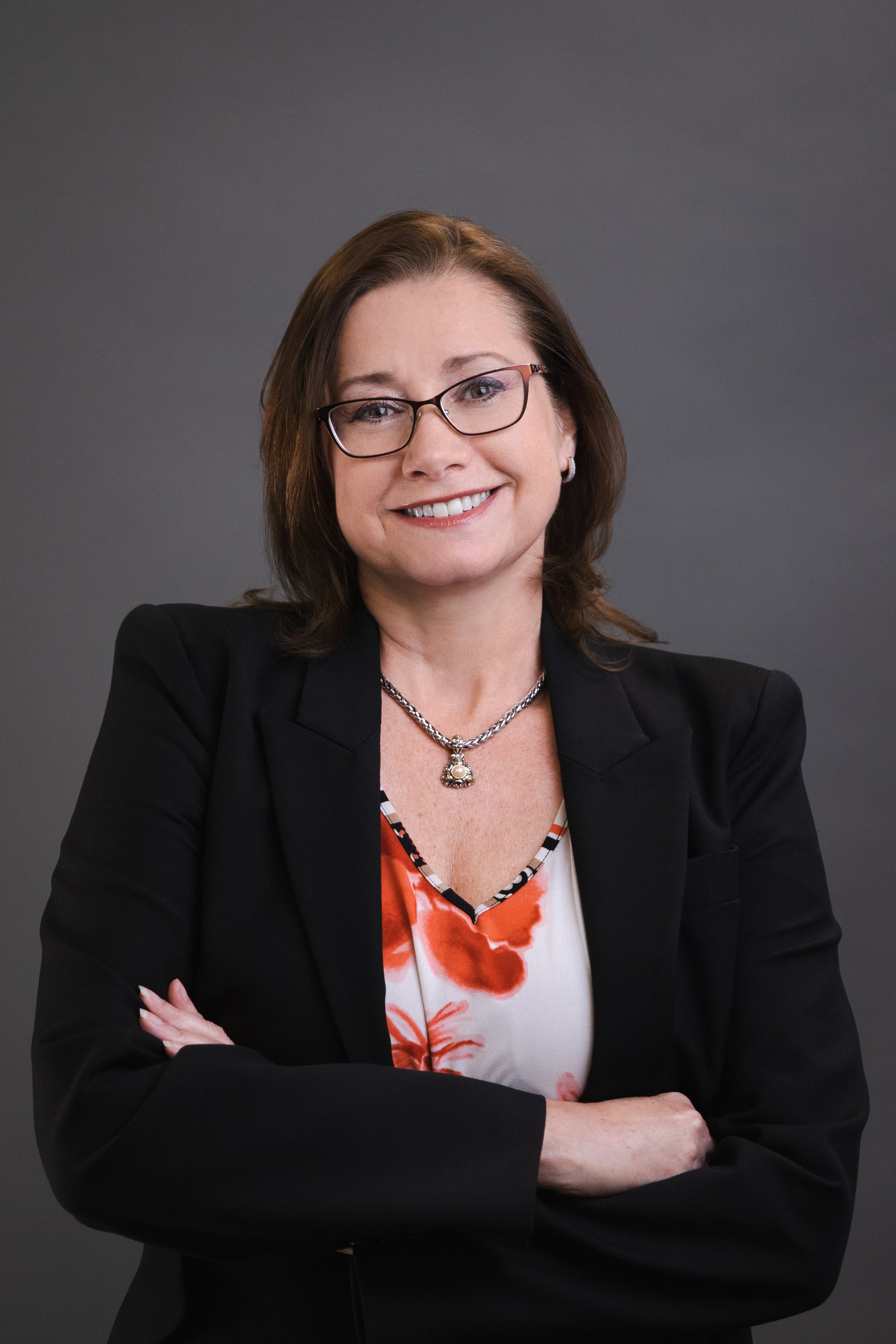 A woman with brown hair, glasses, and a smile, wearing a black blazer, a colorful blouse with orange and white floral pattern, a necklace with a pendant, and crossed arms, standing against a gray background.