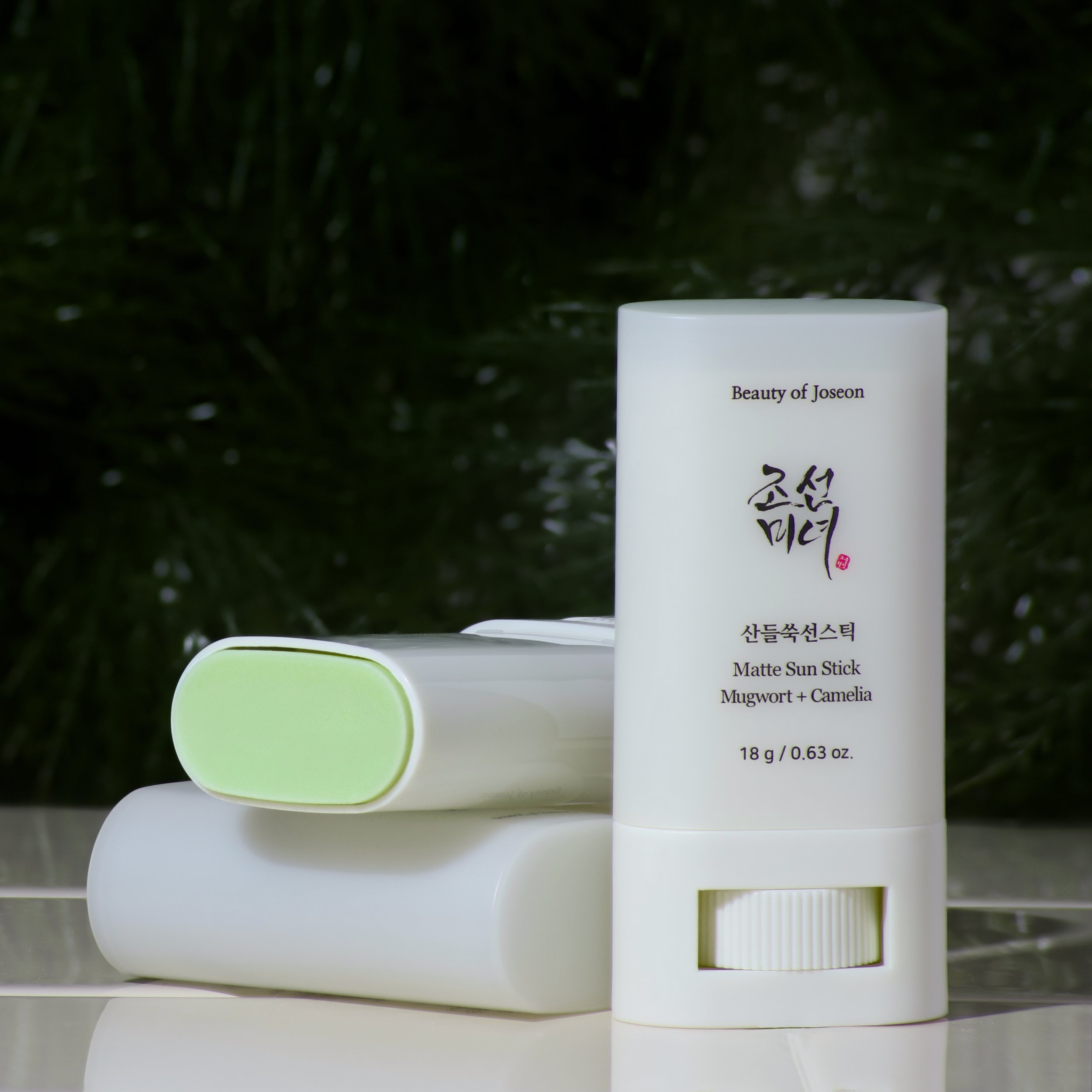 A pale green cylindrical container of a Beauty of Joseon sunscreen stick, placed on a beige tiled surface, with an opened container of it behind showing the bright green product. The background is dark green plant foliage.