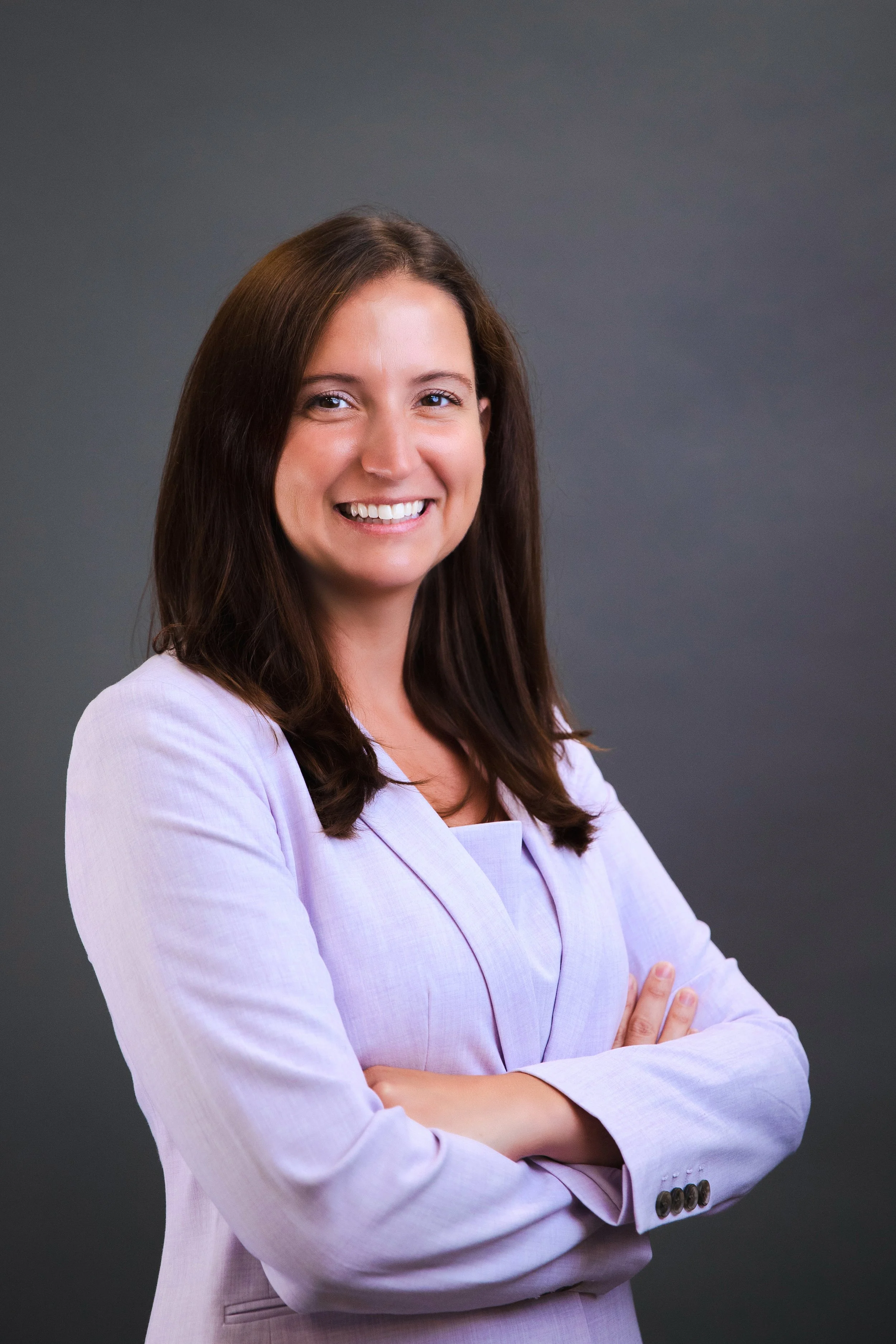 A woman with brown hair smiling, wearing a light purple blazer, standing against a dark grey background with her arms crossed.