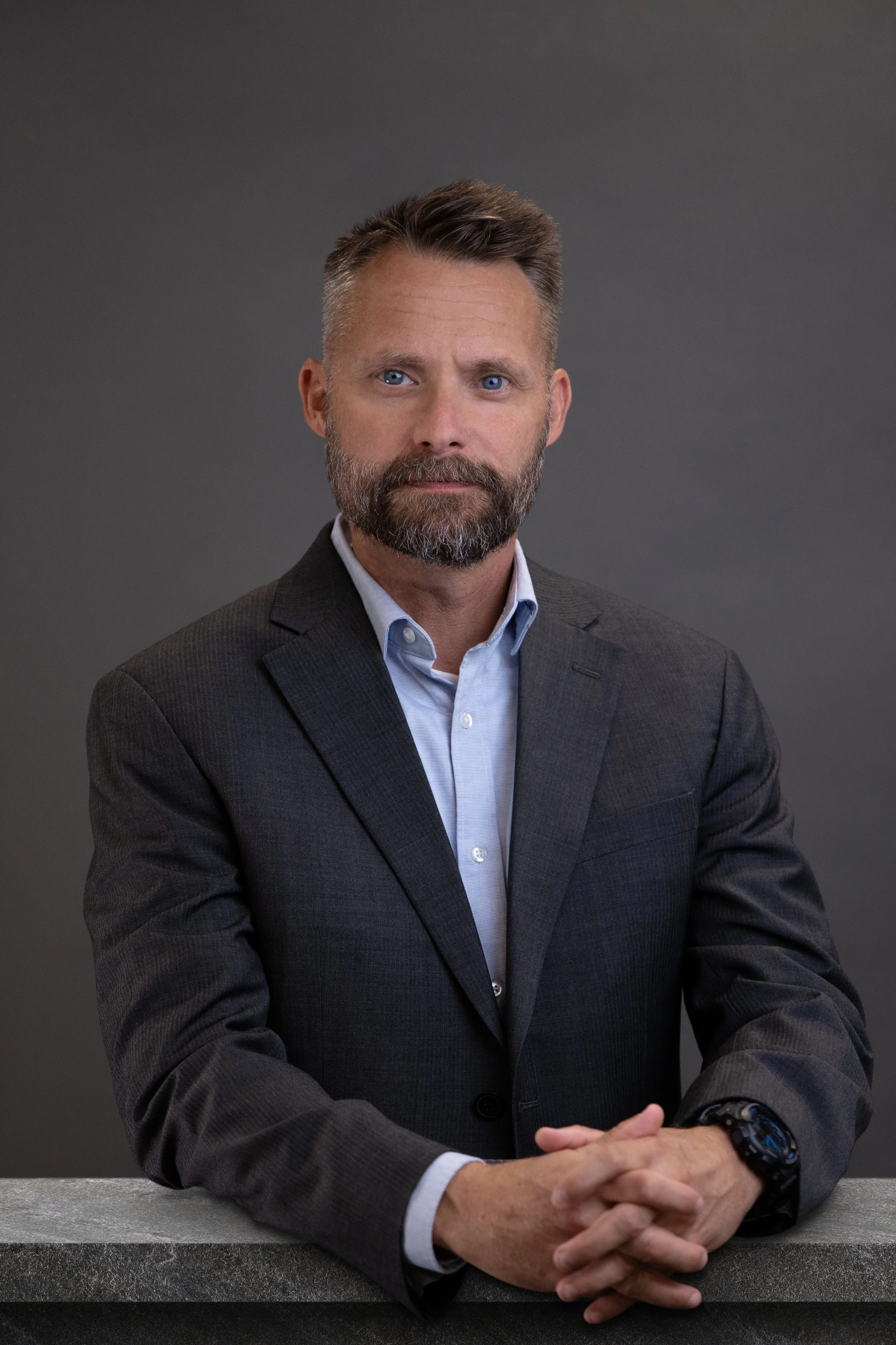 A man with a beard and blue eyes wearing a gray suit and white shirt sitting at a table with hands clasped.