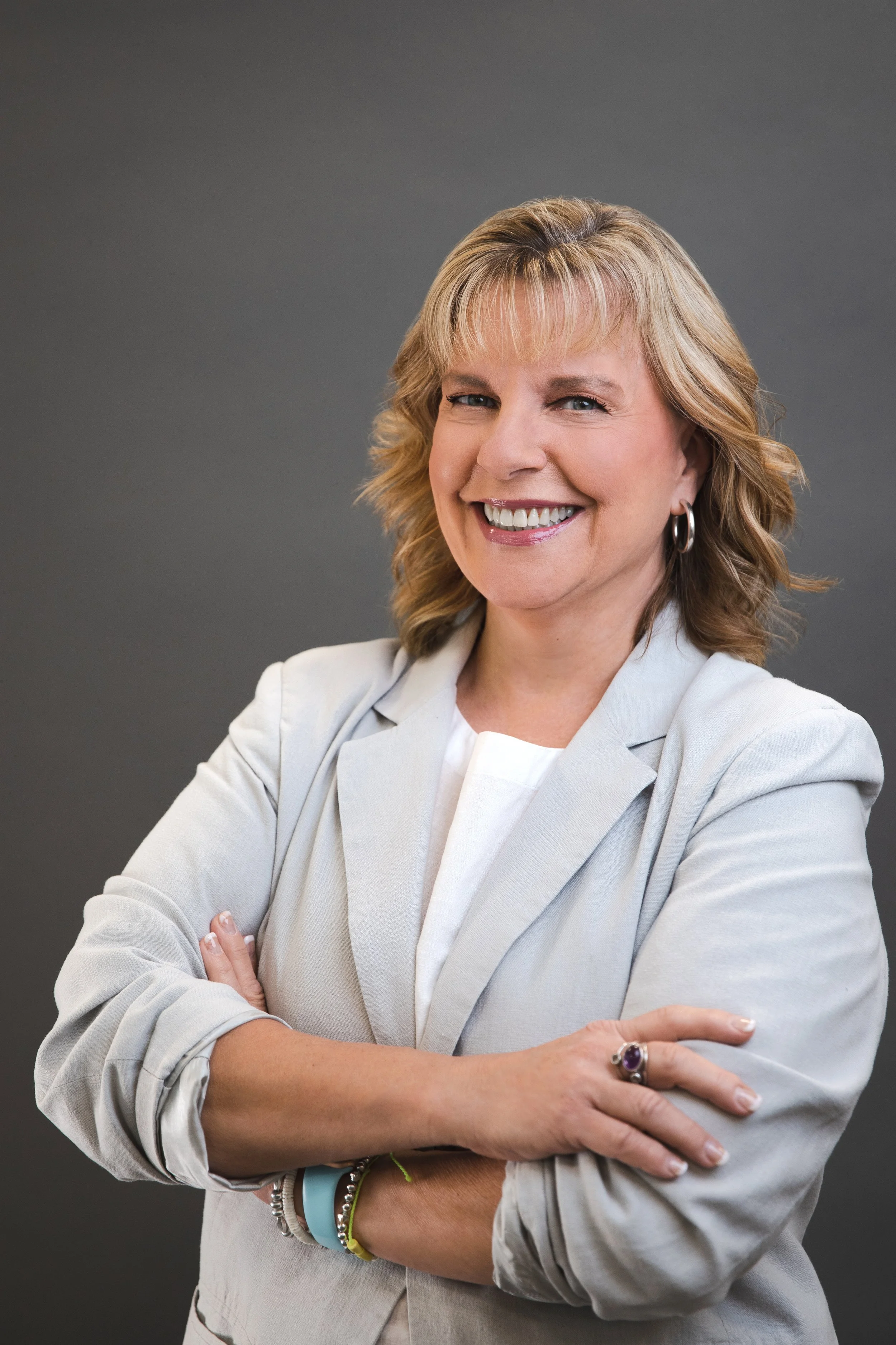 A woman with blonde hair smiling and crossing her arms, wearing a white blazer, earrings, and colorful bracelets, against a dark gray background.