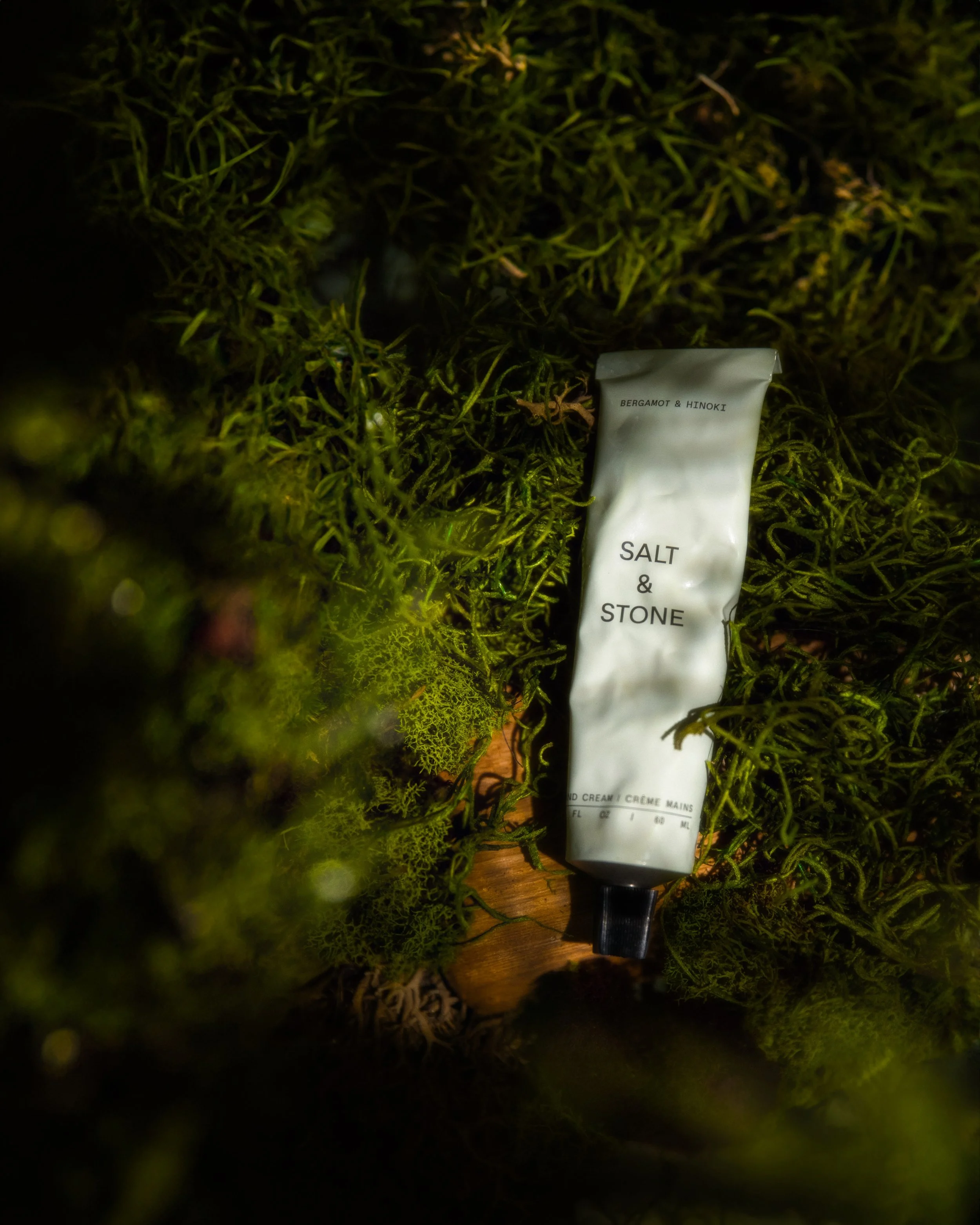 Close-up of mossy green foliage with a sage tube of hand cream labeled 'Salt & Stone - Bergamot & Hinoki' lying on the moss and cedar, in a dimly lit forest environment.