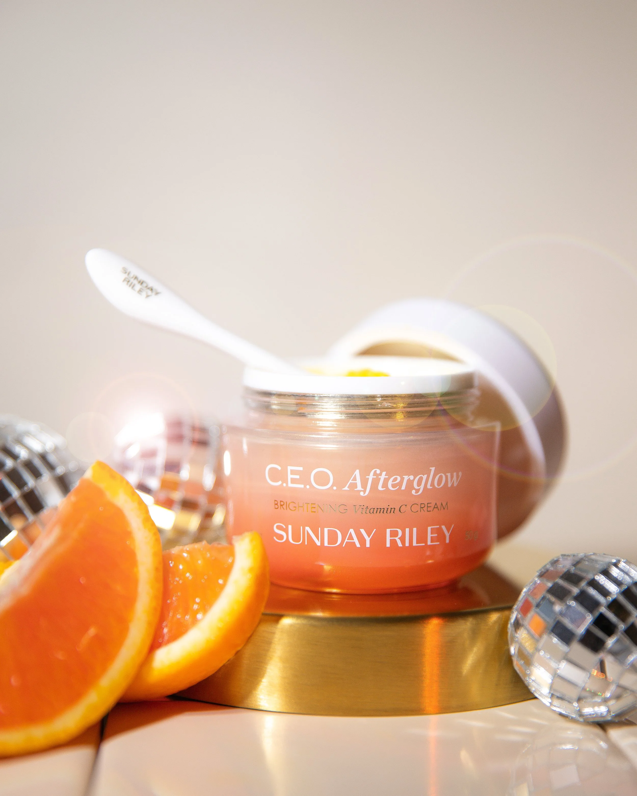 Close-up of a skincare product labeled Sunday Riley C.E.O. Afterglow, Brightening Vitamin C Cream, with sliced orange and a small crystal decorative item nearby, on a golden surface.
