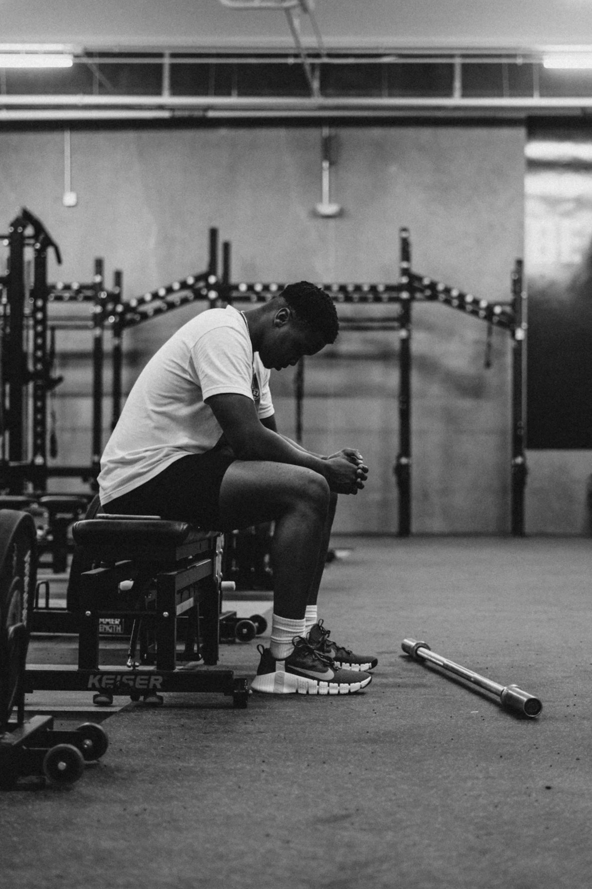 A man sitting on a workout bench in a gym, looking down with his hands clasped, surrounded by fitness equipment including a barbell on the floor, with empty wall-mounted racks in the background.
