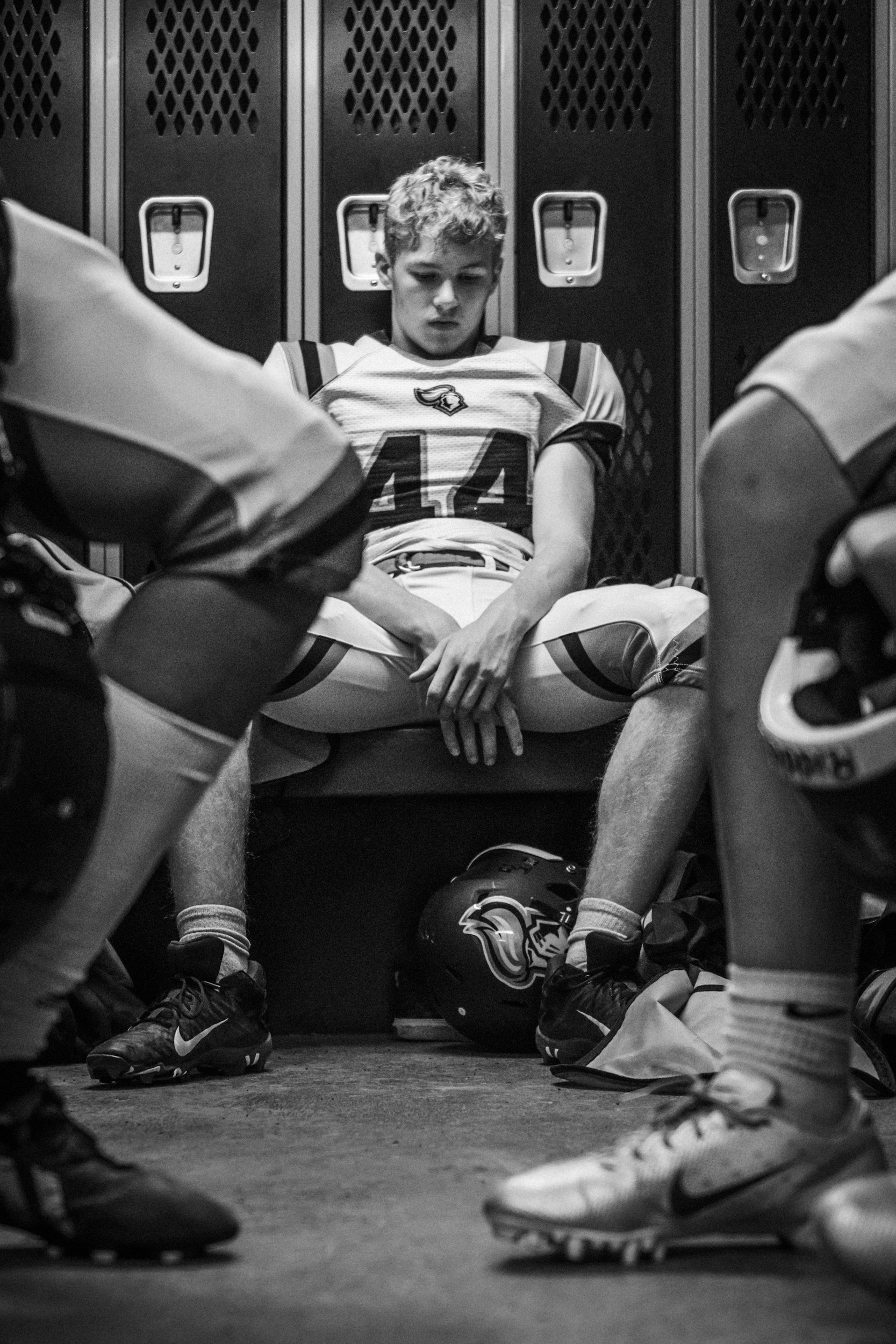 A young football player sitting on a bench in a locker room, looking down with a somber expression. He is in a football uniform with the number 44, surrounded by other players in similar attire. The scene is in black and white.
