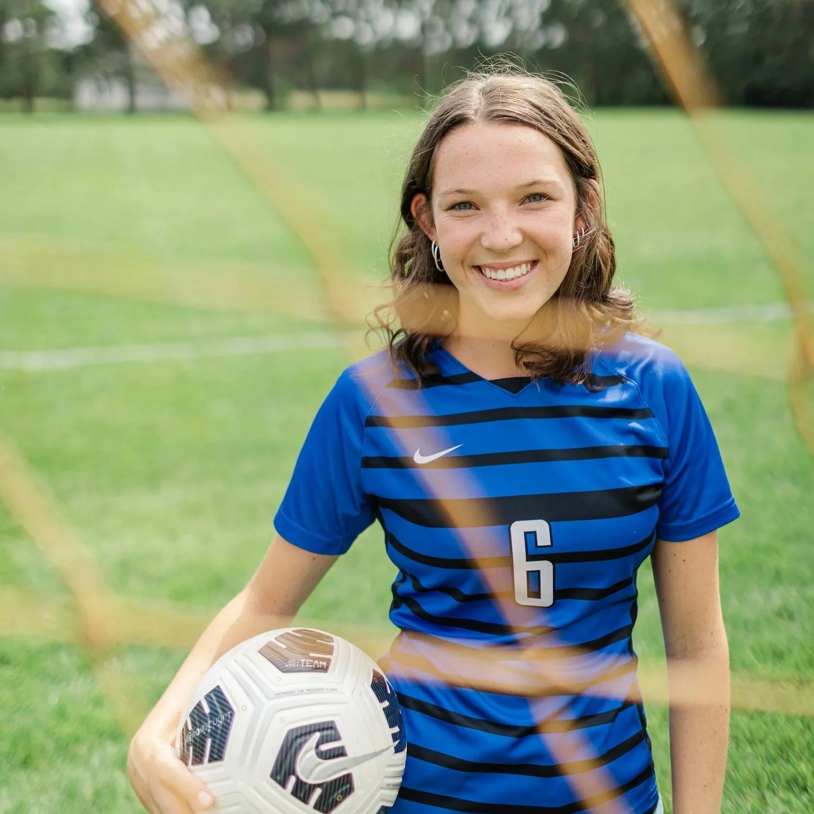 A young woman smiling, wearing a blue soccer jersey with the number 6, holding a white soccer ball with black and colored patterns on a grassy field surrounded by trees.